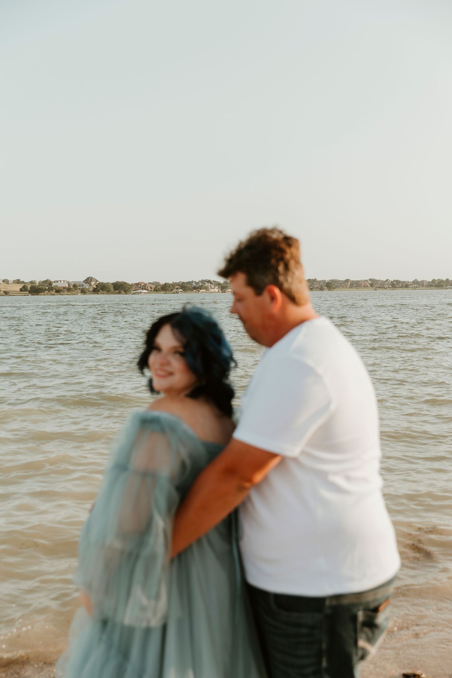A couple shares an emotional embrace on a beach with water and shoreline in the background.