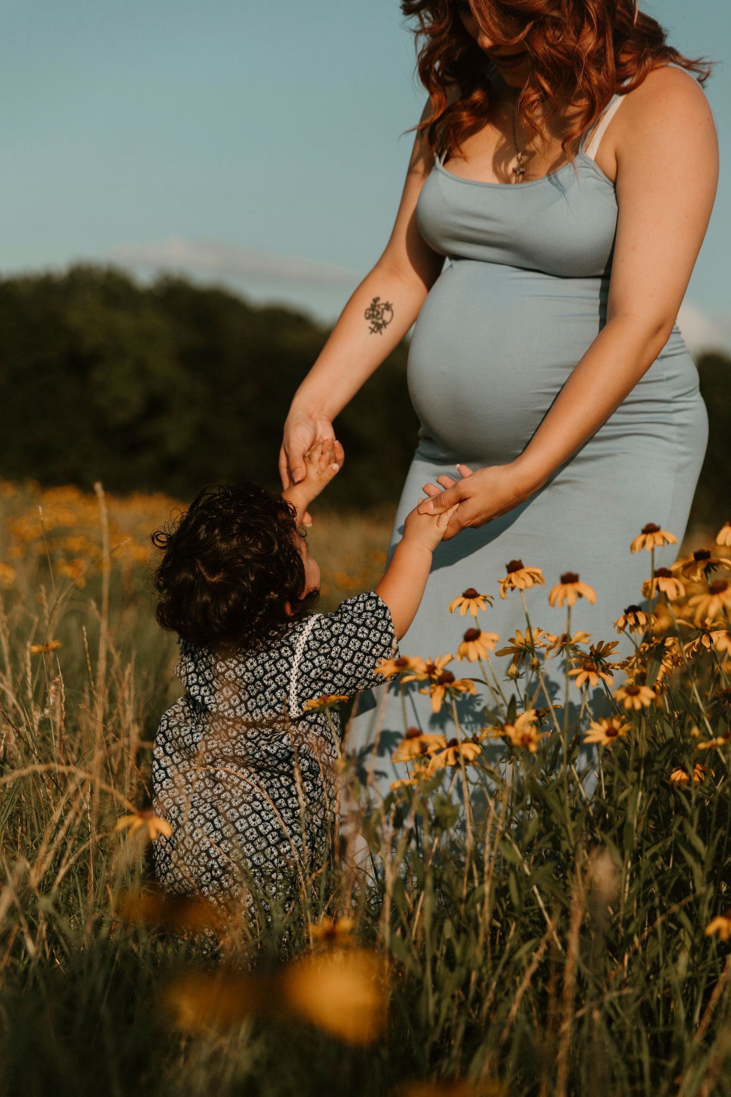 Close-up view of maternity dress and floral print outfit against yellow wildflowers at sunset.