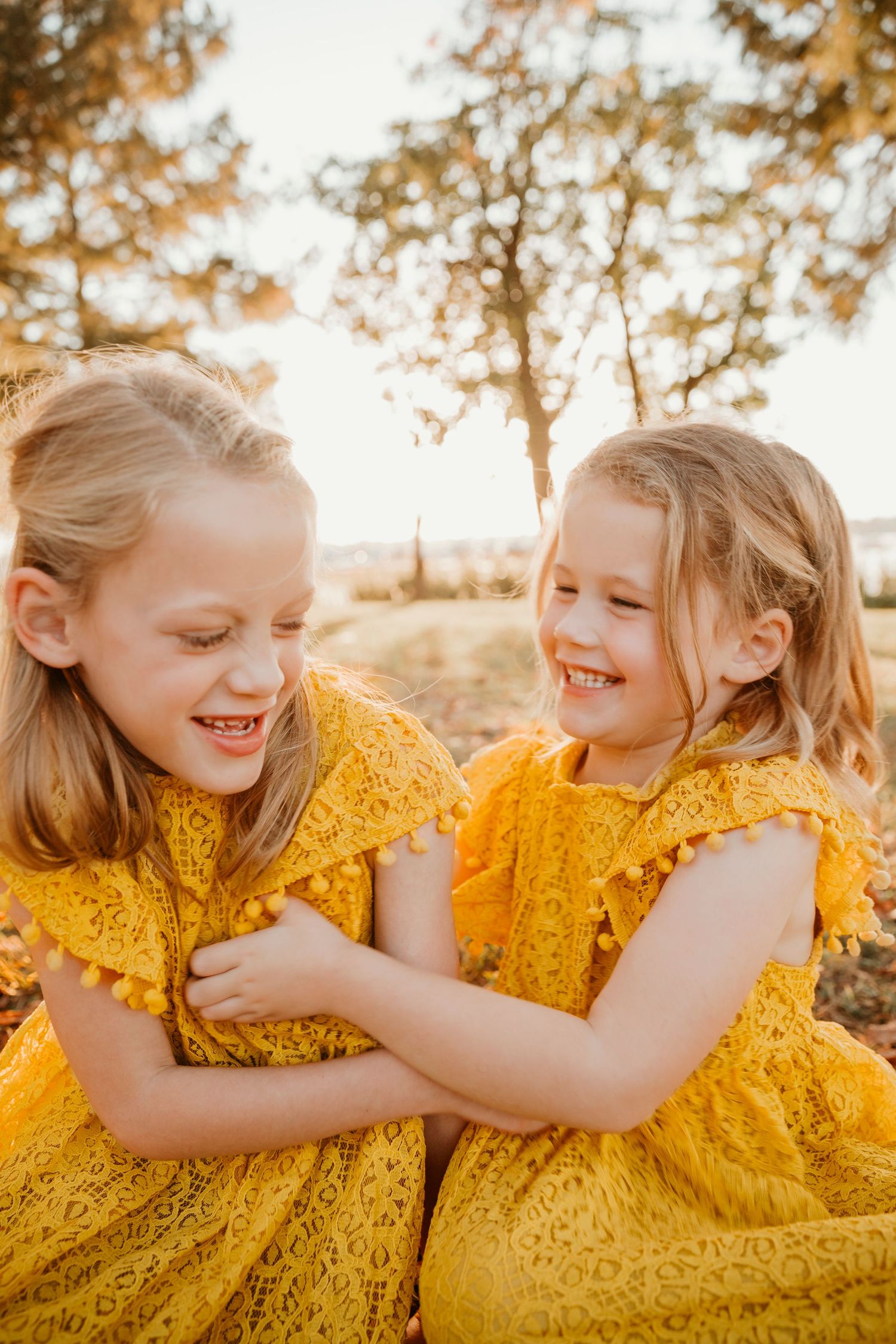 Two young girls in matching yellow lace dresses laugh and embrace during a golden sunset photo session.