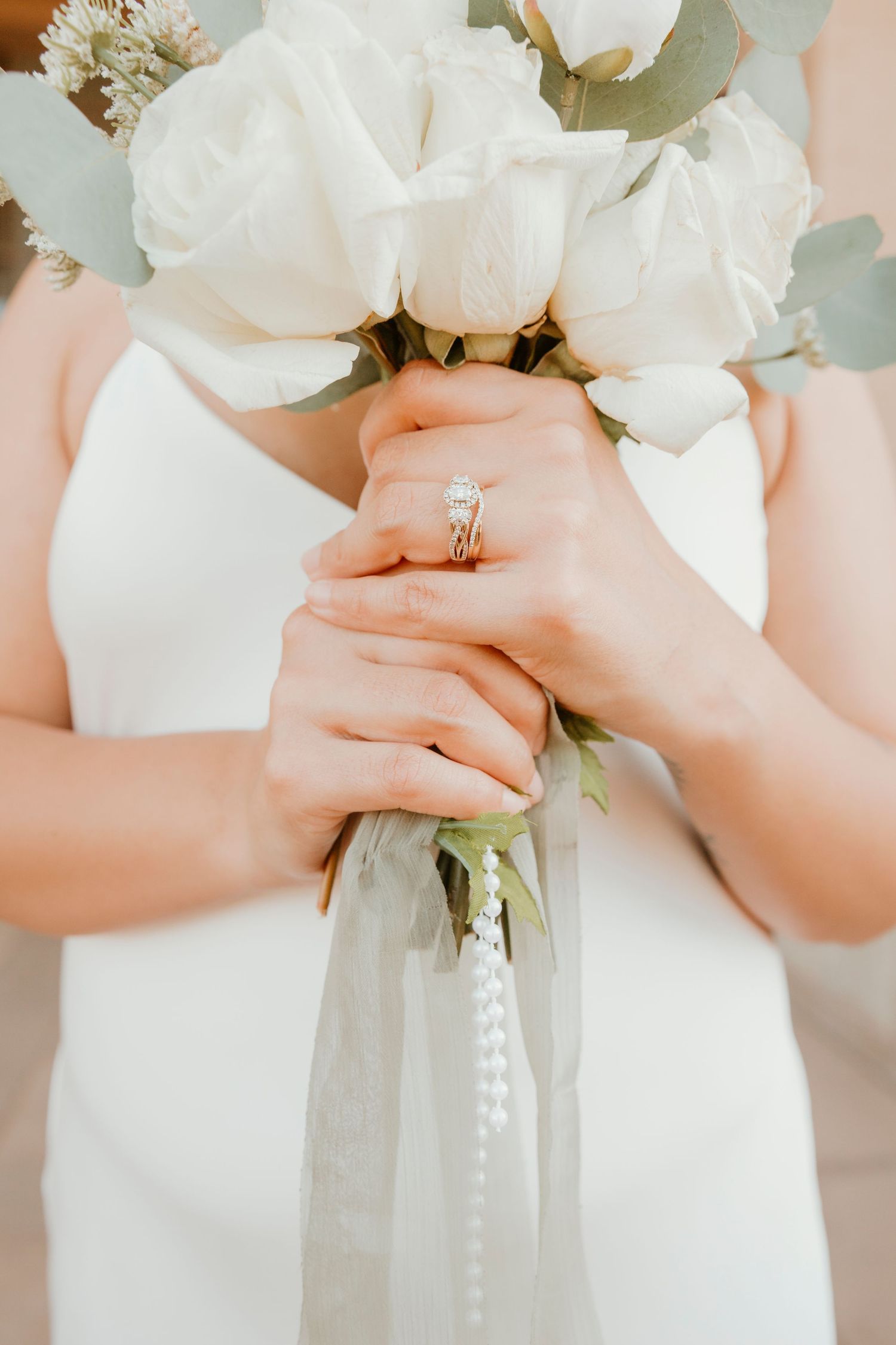 Close-up view of hands holding an elegant white bridal bouquet with eucalyptus leaves against a soft pink background.