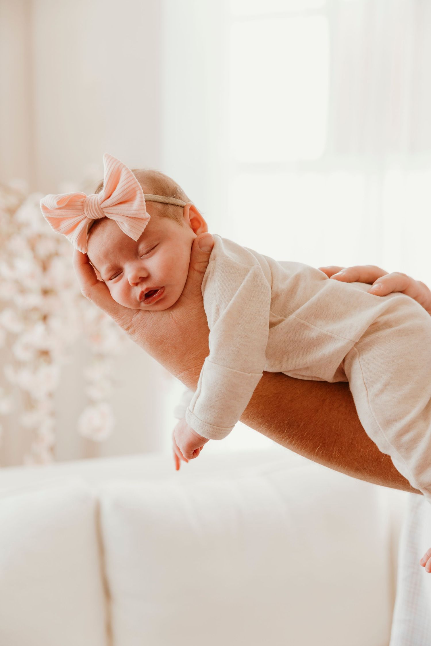 Sleeping newborn wearing white onesie and pink bow headband being held against white background.