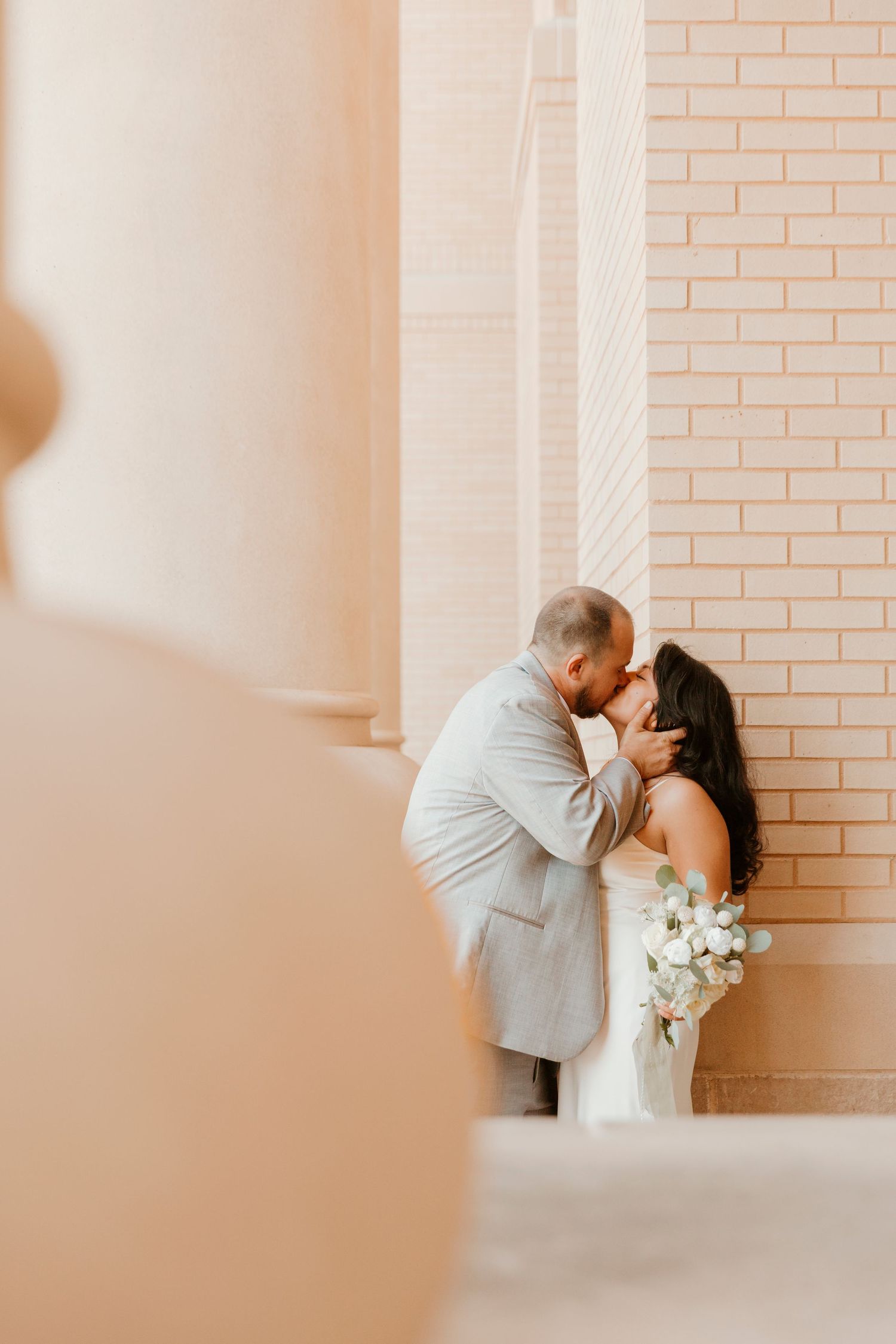 A romantic couple in light gray and white attire share a tender kiss against a blush pink brick wall.