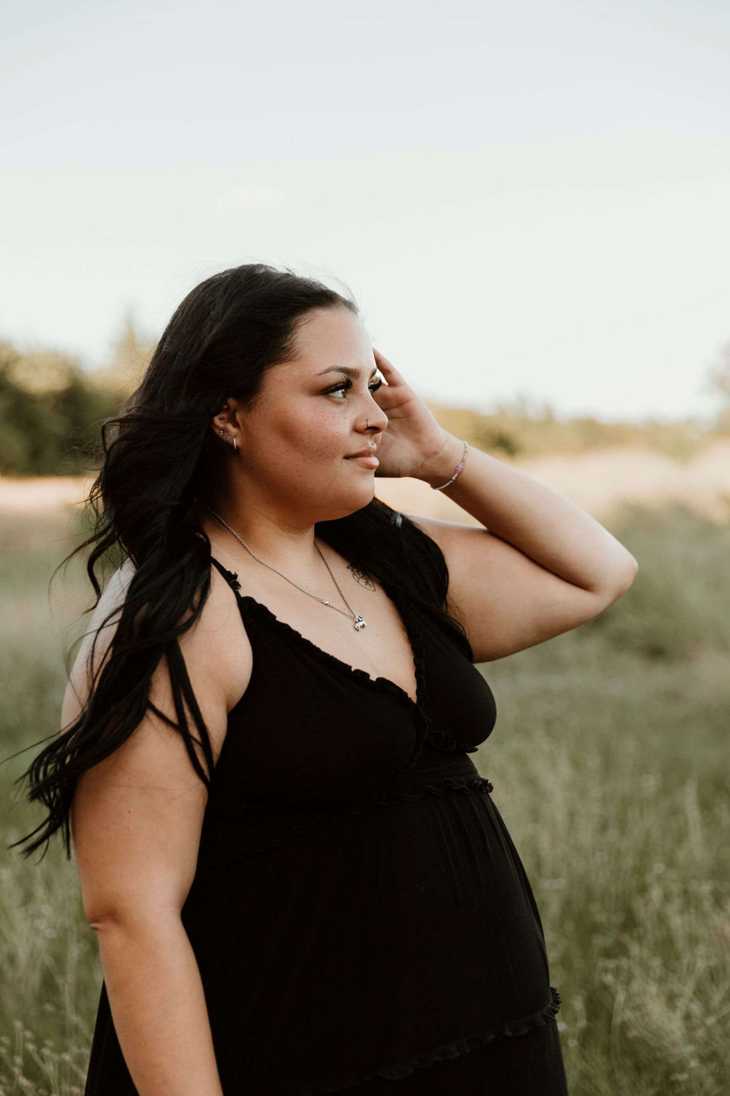 A woman in a black dress stands in a field at sunset, running her hand through her long dark hair.