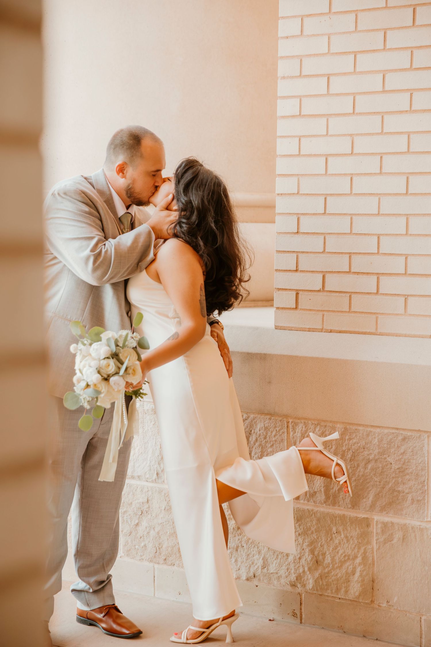 A romantic couple in wedding attire share a tender kiss against a brick wall while holding a white floral bouquet.