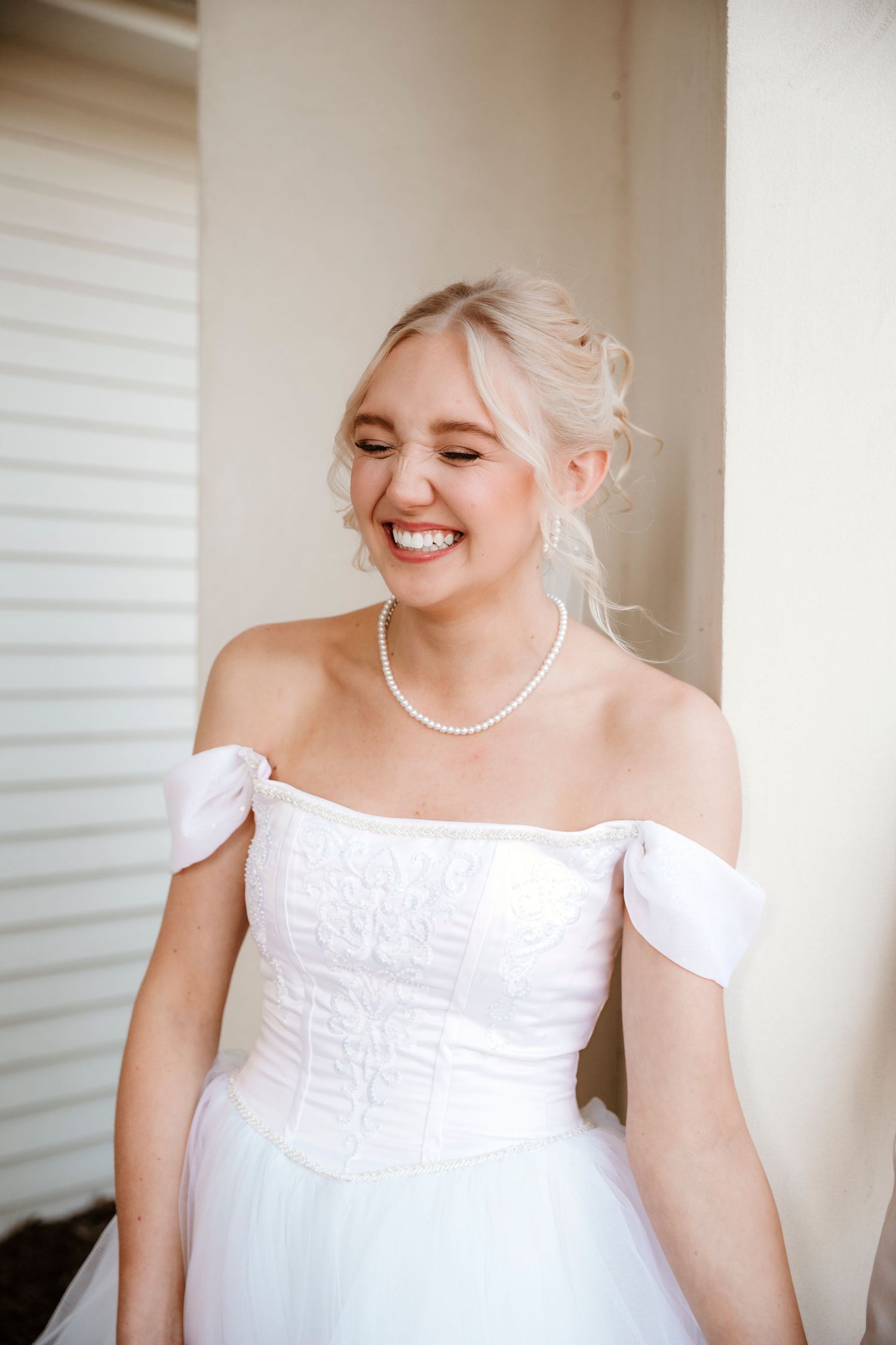 A bride in a white off-shoulder dress and pearl necklace smiles joyfully during her wedding day.