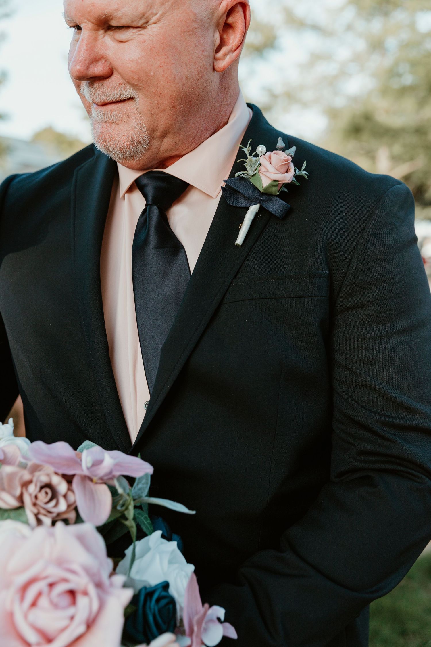 Close up detail of black tuxedo with pink boutonniere and matching pink floral bouquet at formal wedding ceremony.