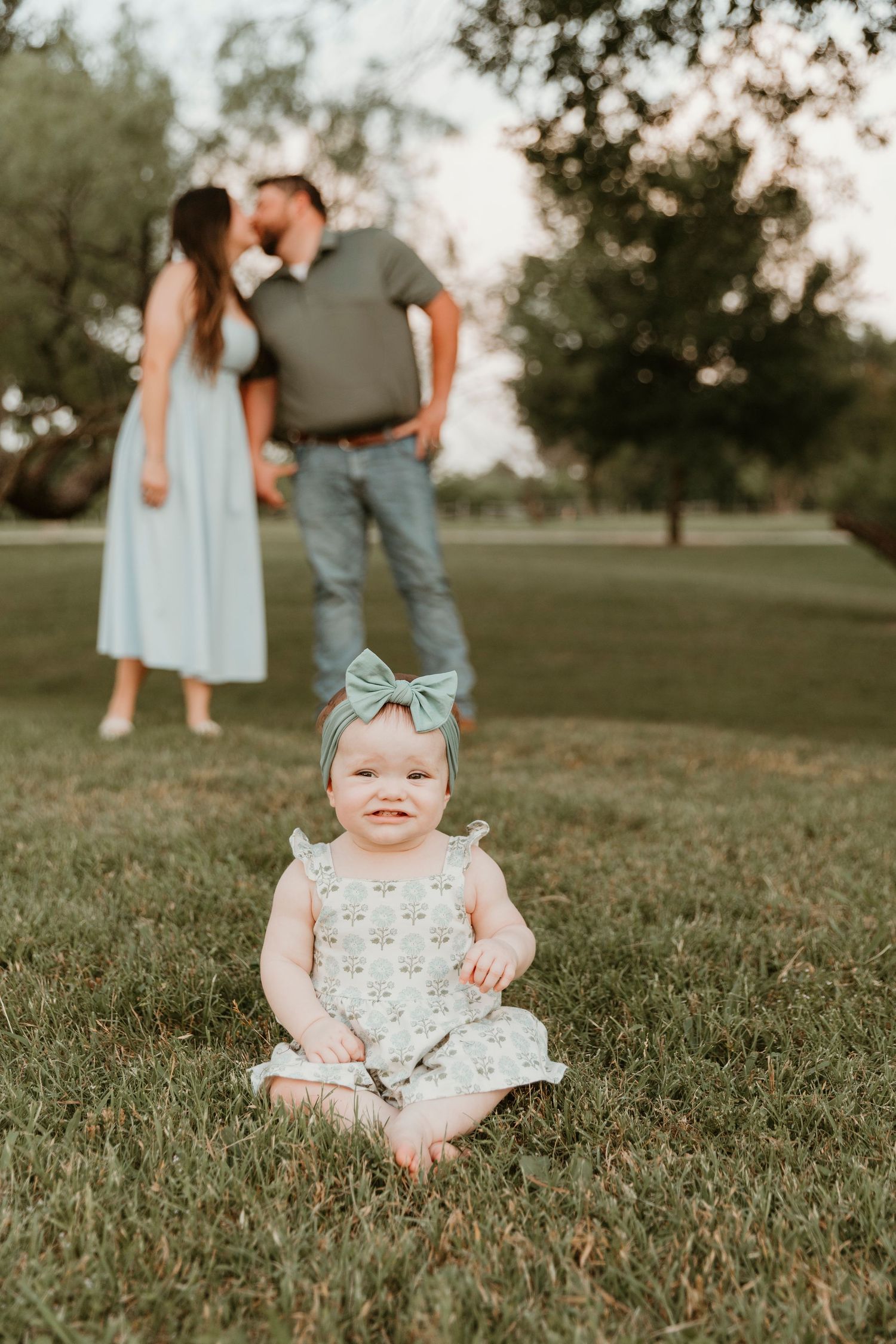 A baby in floral outfit and teal bow sits on grass while two people stand in background.