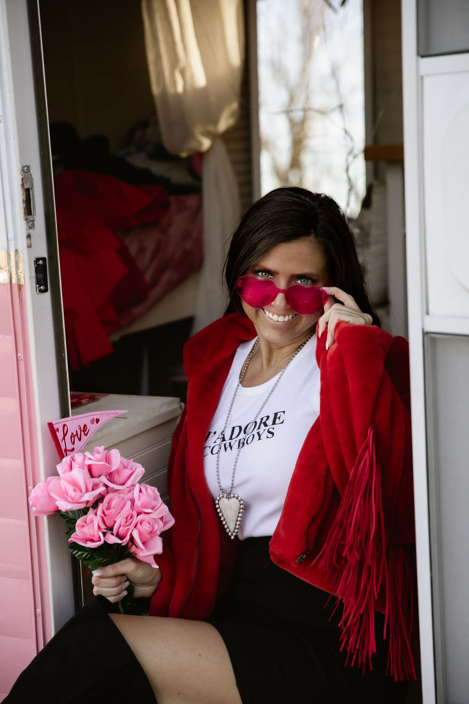 Woman in red cardigan and pink sunglasses poses with pink flowers in doorway.