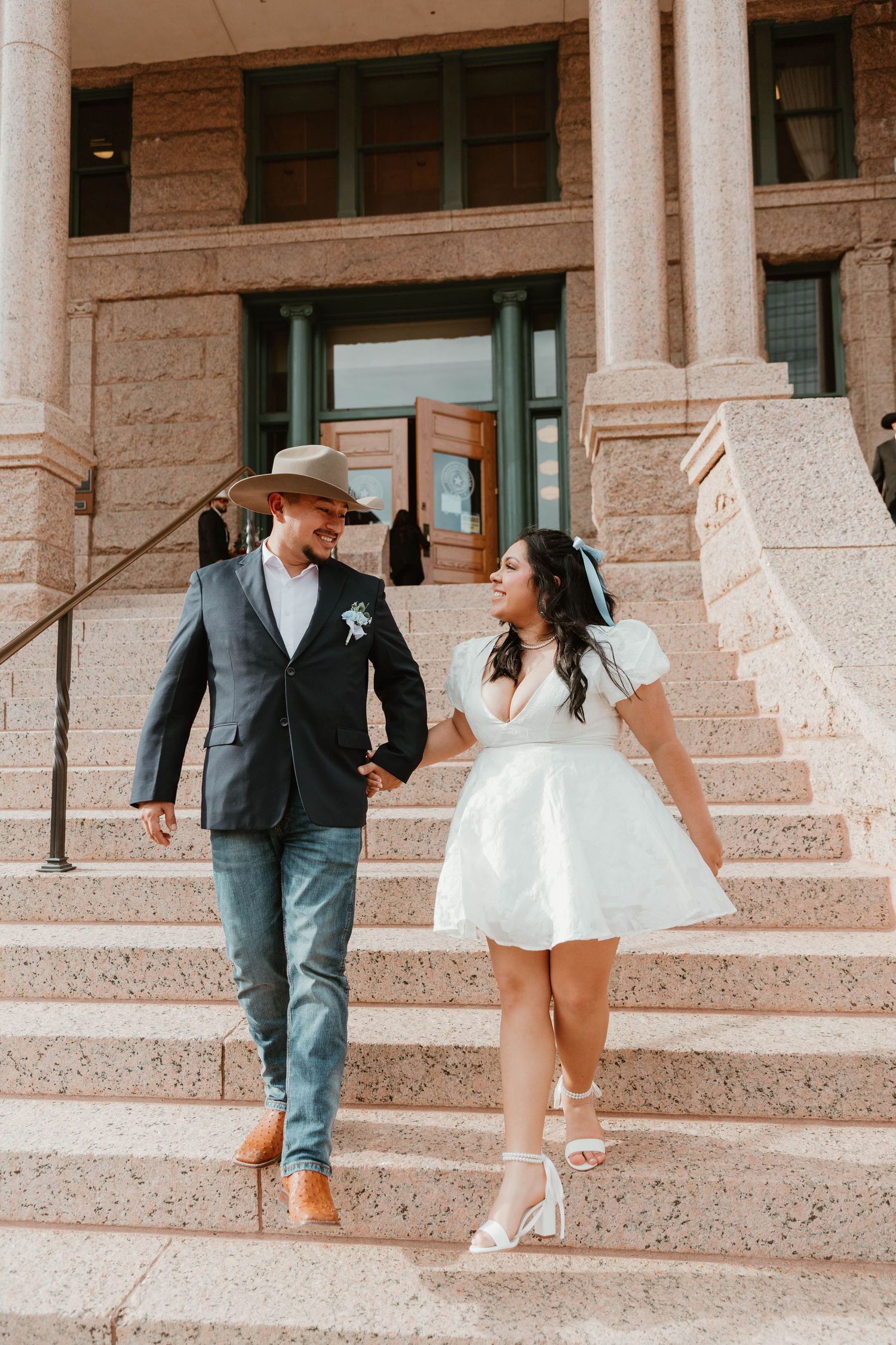 Couple descending stone courthouse steps in wedding attire on sunny day.