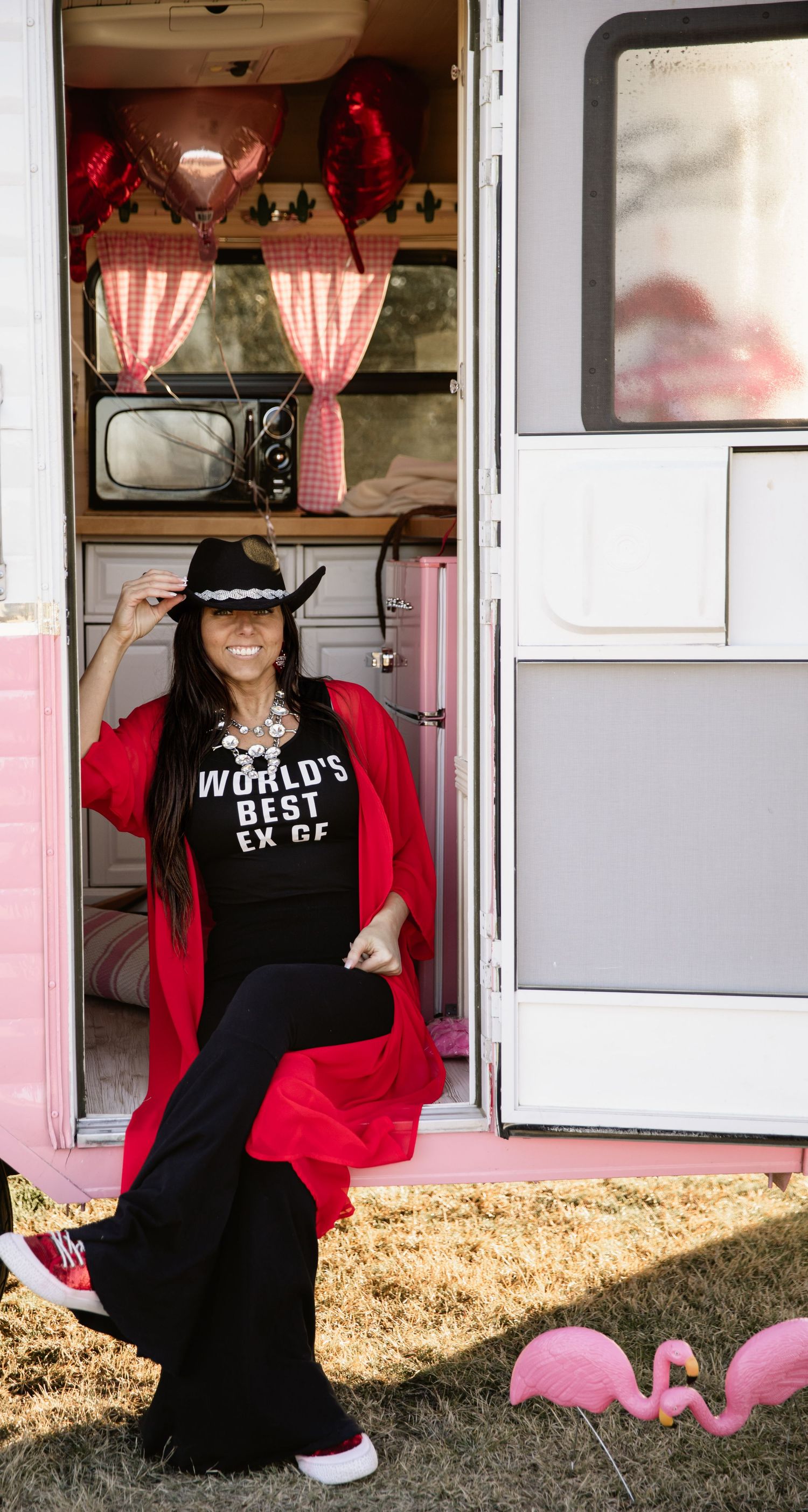 Person in cowboy hat and red cardigan poses in vintage camper doorway with pink decor.