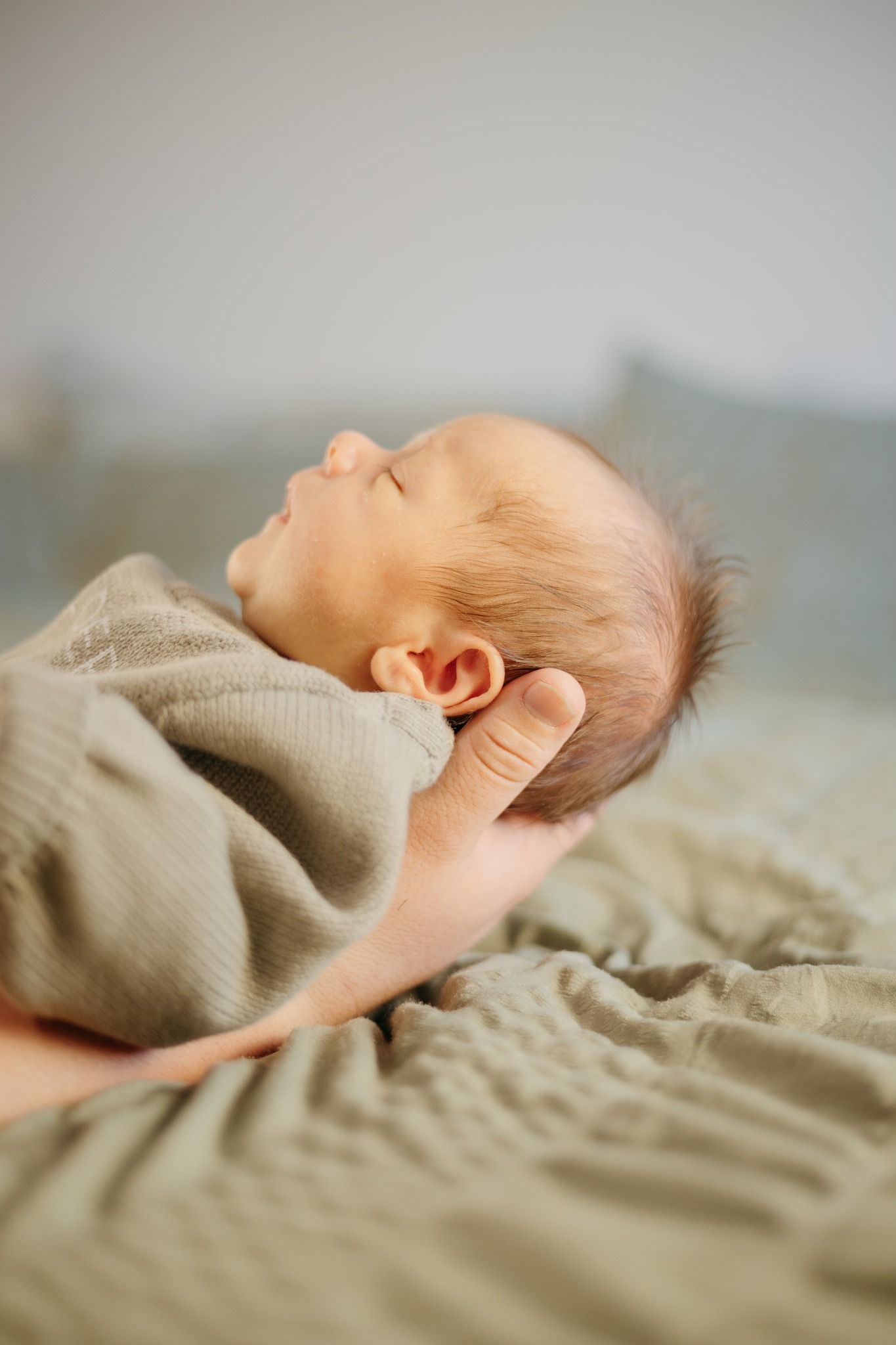 Sleeping newborn with fine hair wearing beige sweater resting on textured bedding.