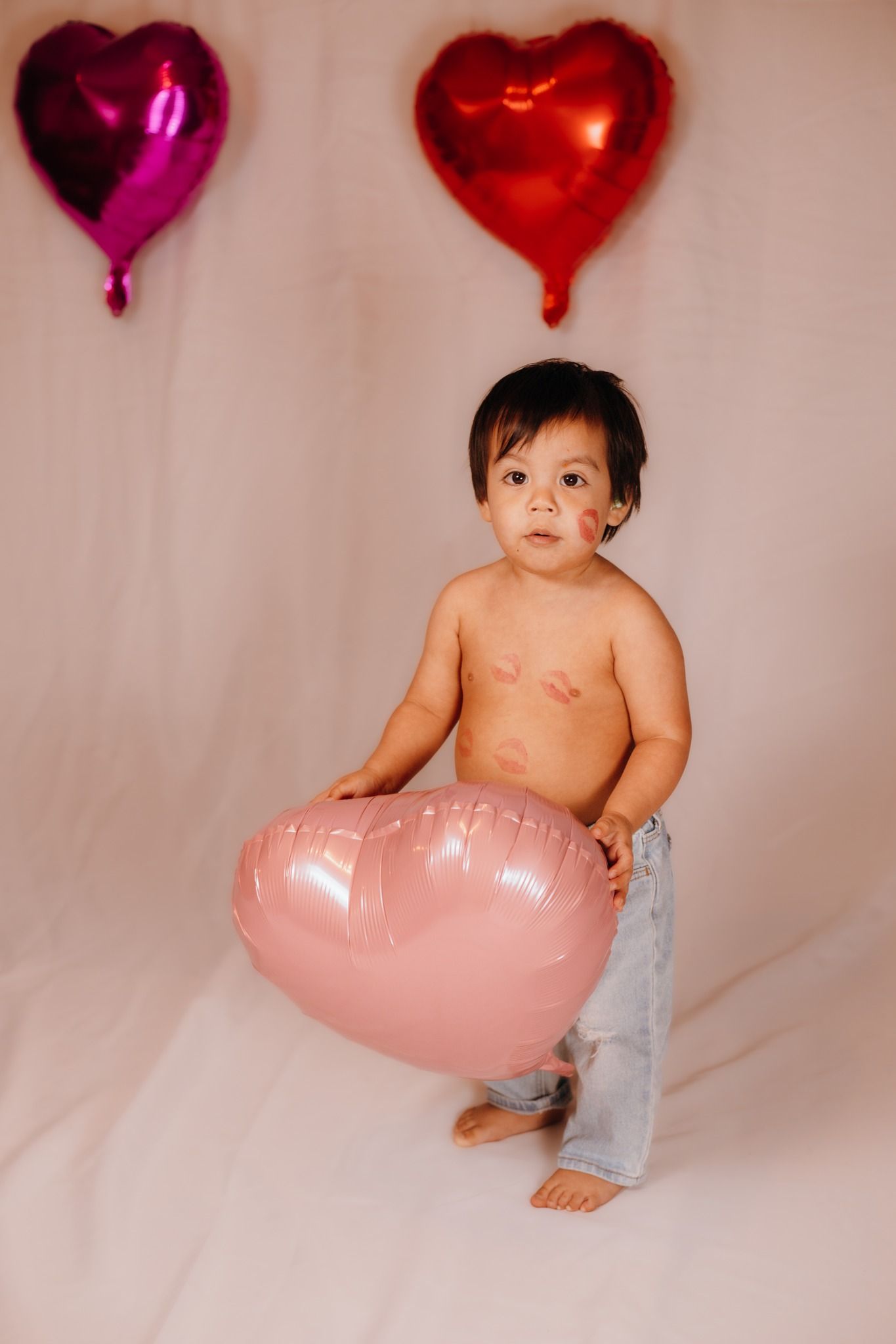 Baby sitting with heart-shaped balloons in pink and red for Valentine's themed photoshoot.