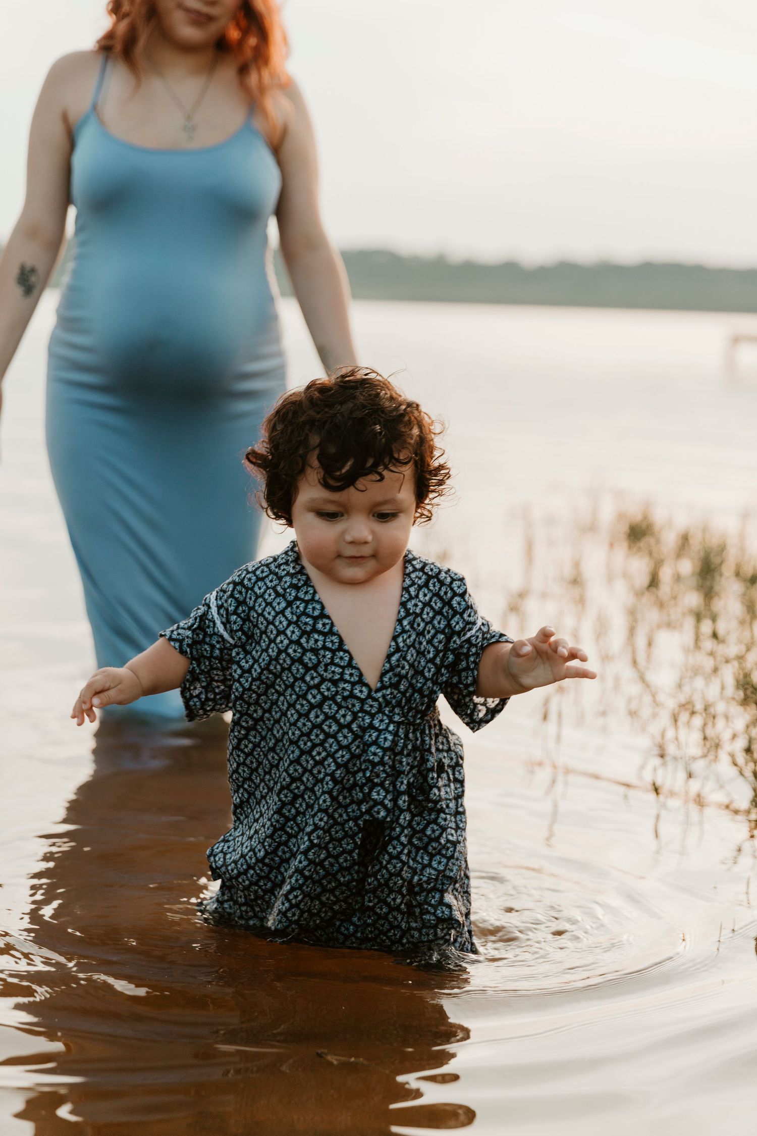 Small child in patterned dress splashes in lake water while supervised.