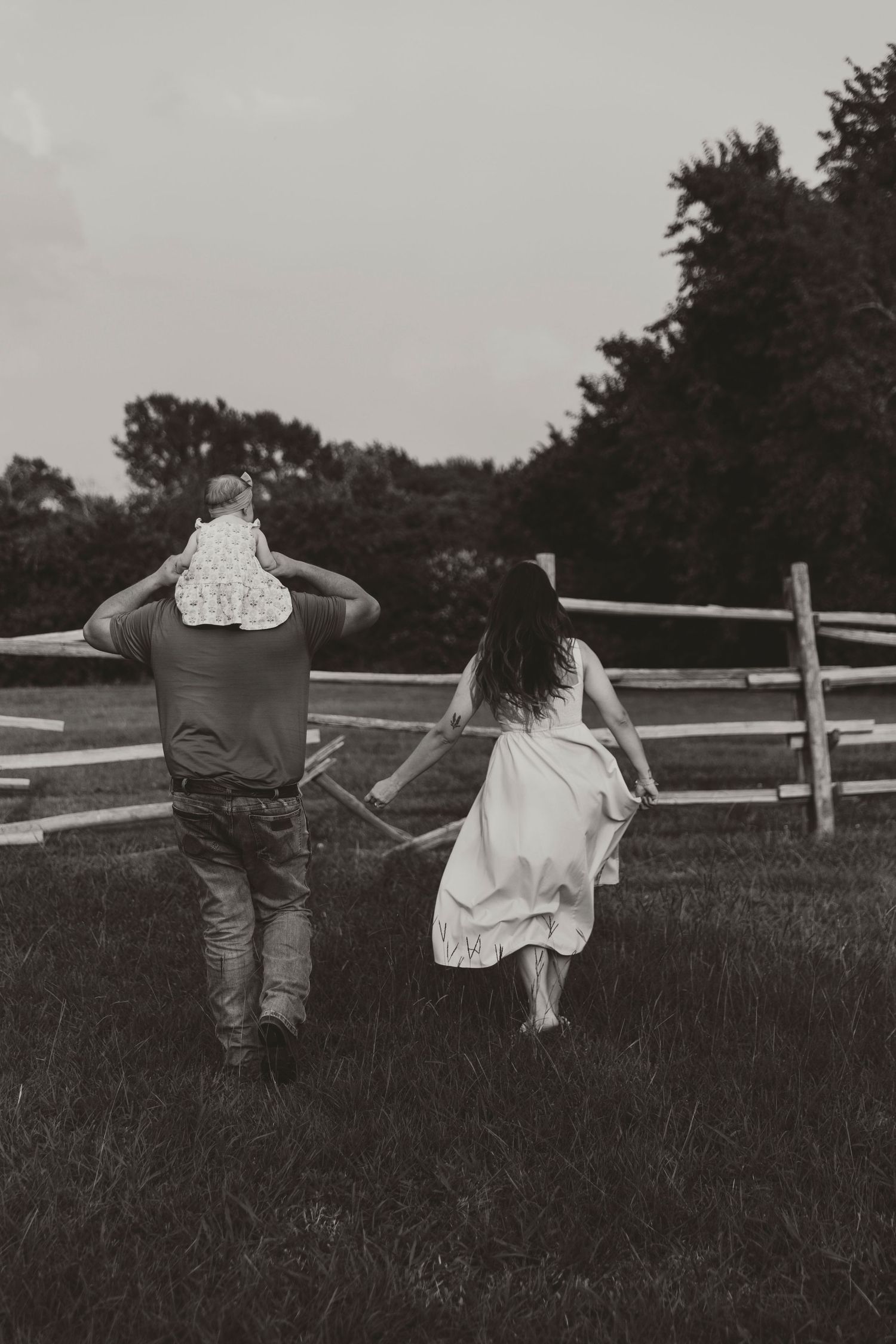 Two people hold hands while running through a field near a rustic fence at sunset.