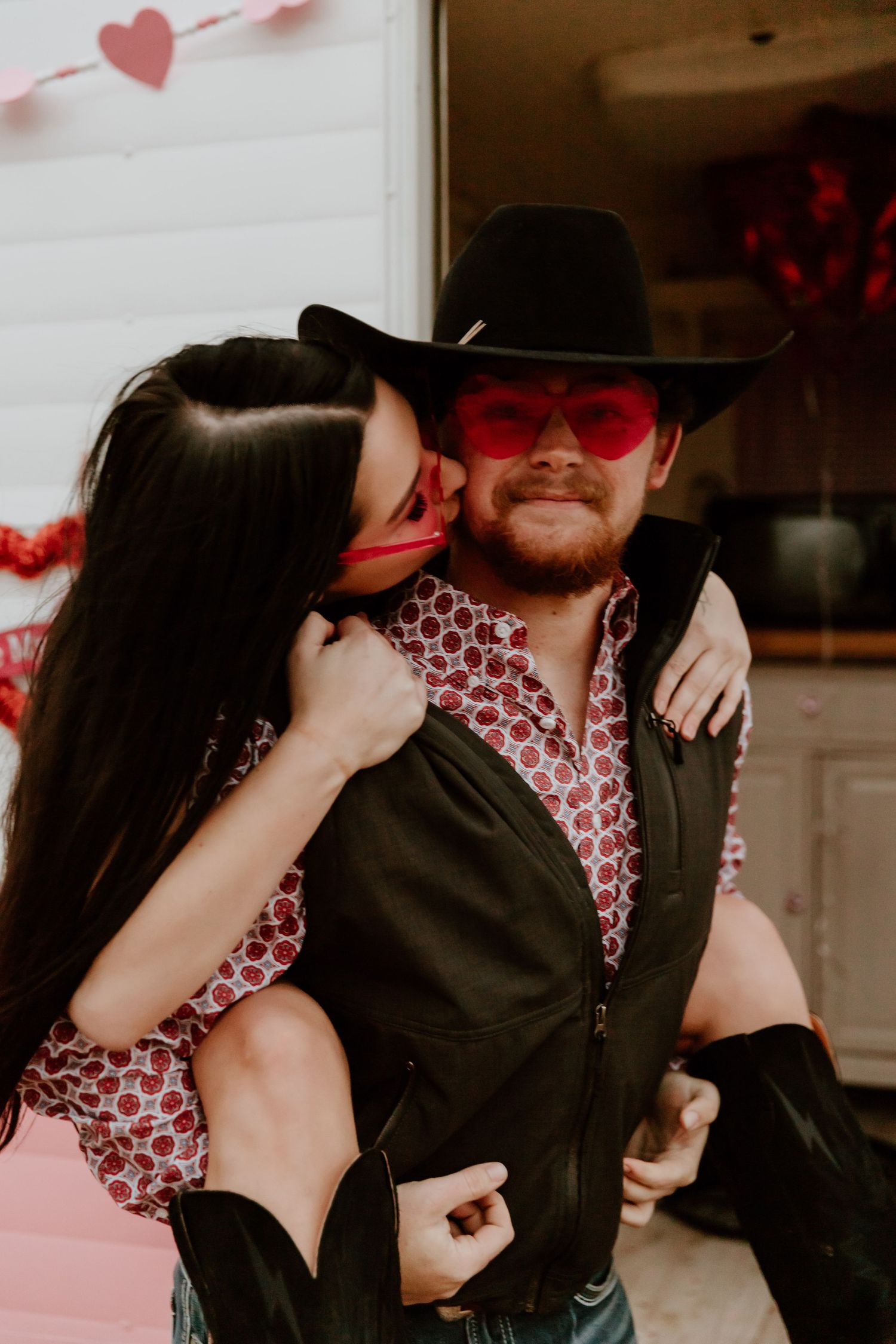 Couple in western-inspired outfits sharing intimate moment with pink Valentine's decor in background.