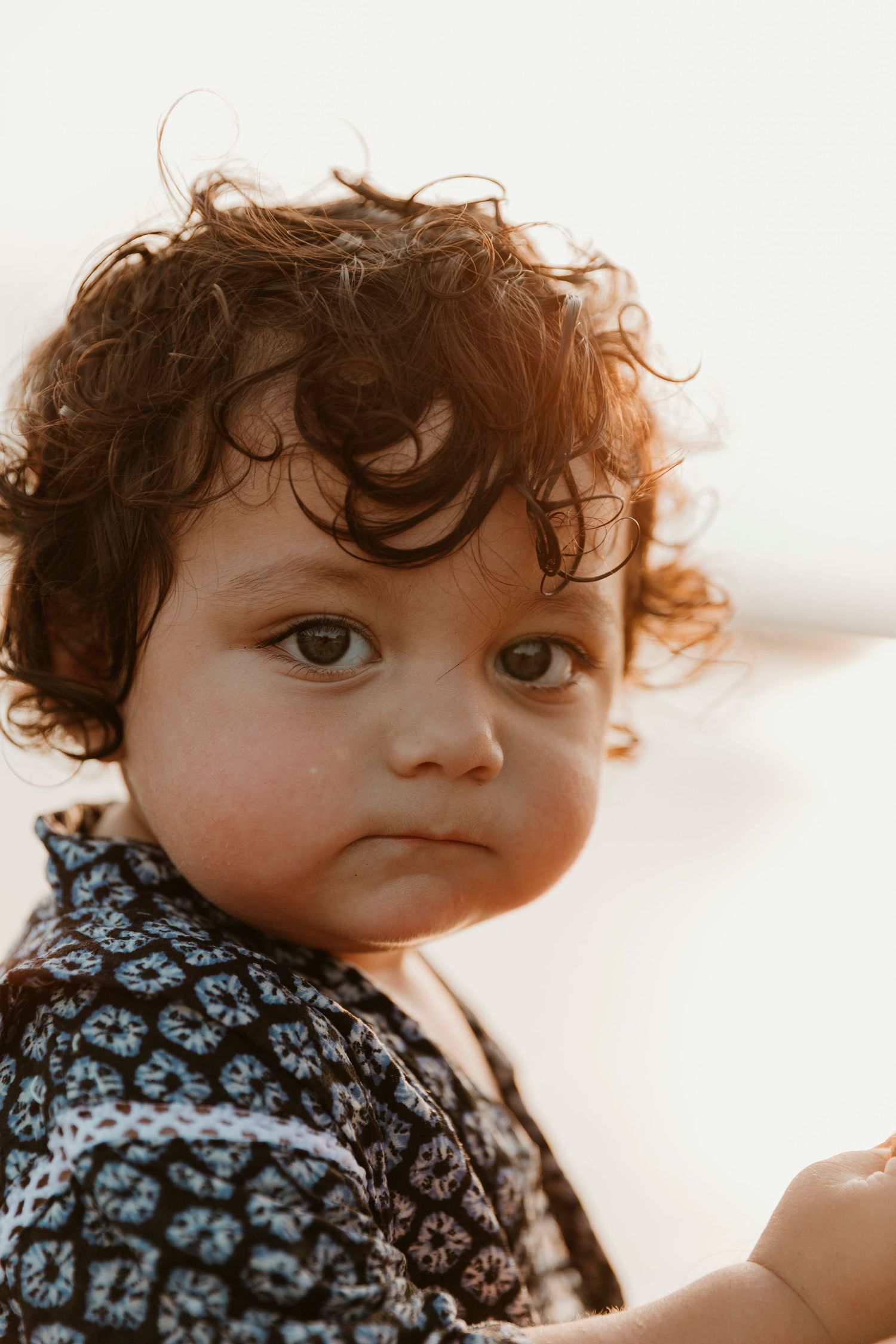 Close-up portrait of a toddler with curly brown hair wearing a patterned shirt against a light background.