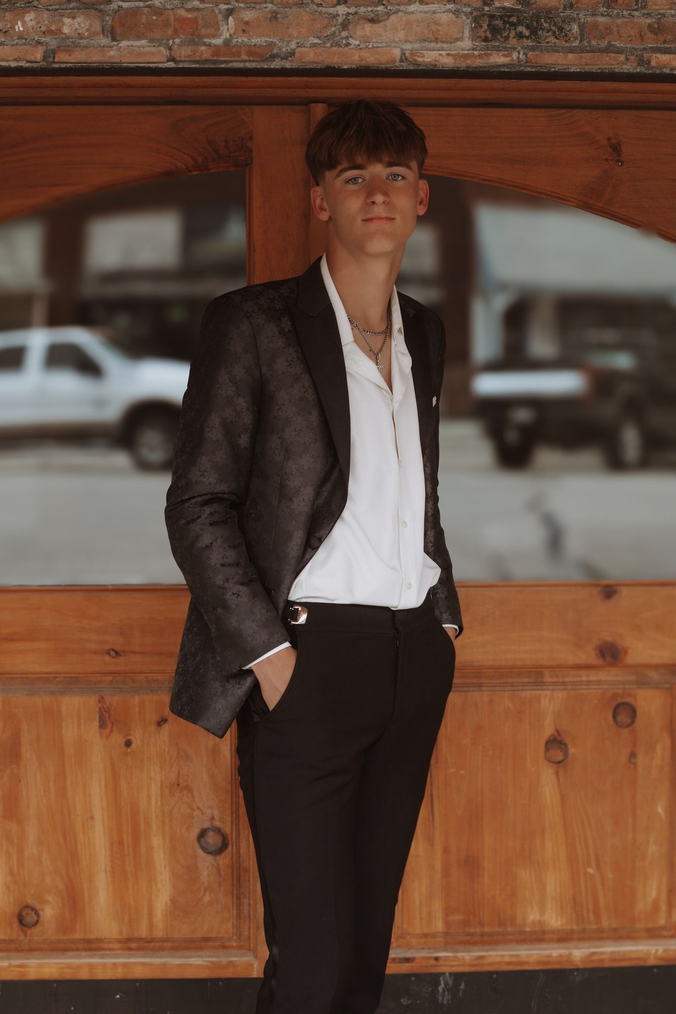 Person in formal black suit and white shirt leaning against wooden paneling.