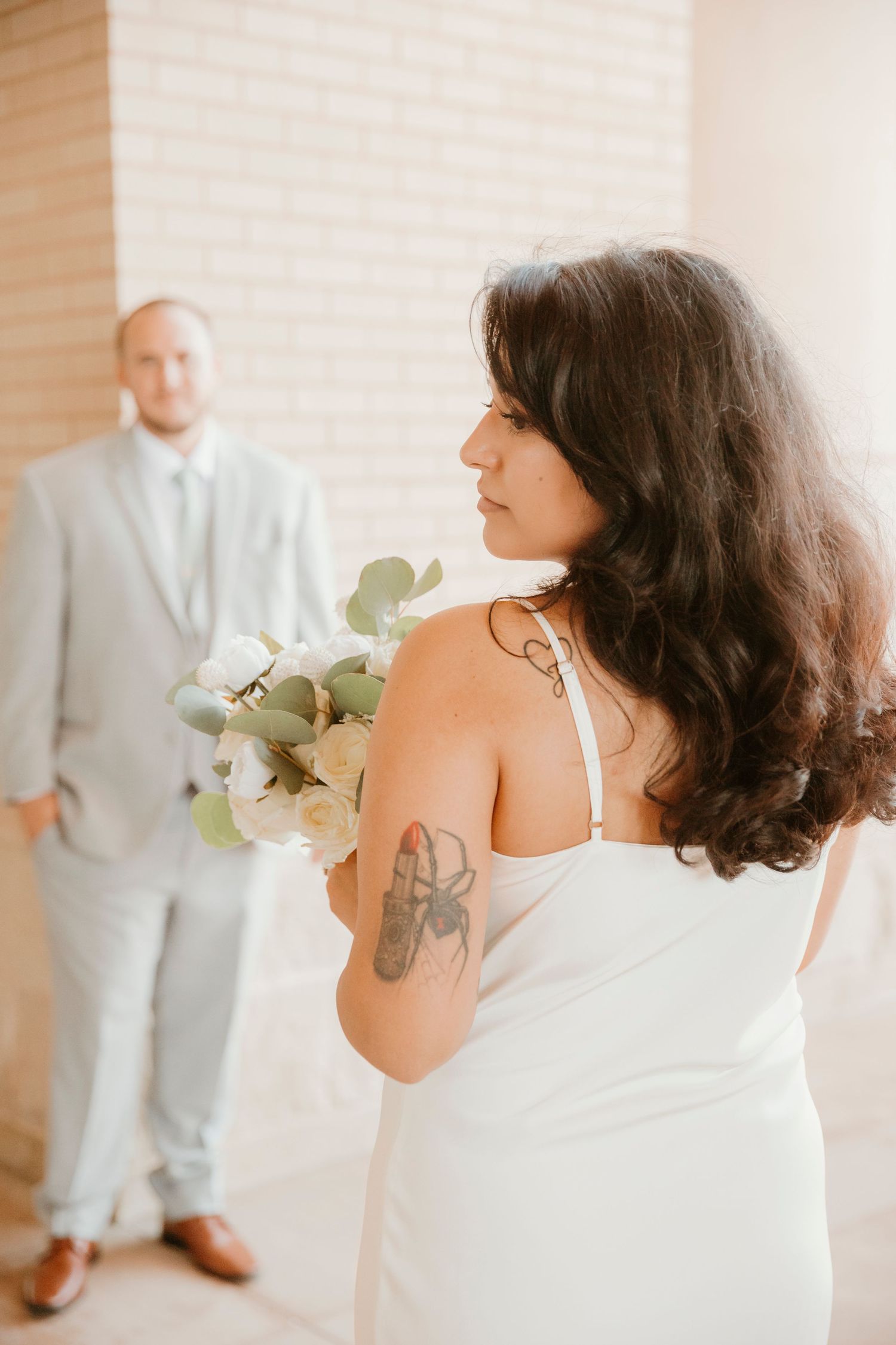 A bride in a white dress with dark hair and tattoos stands in front of a window with natural light.