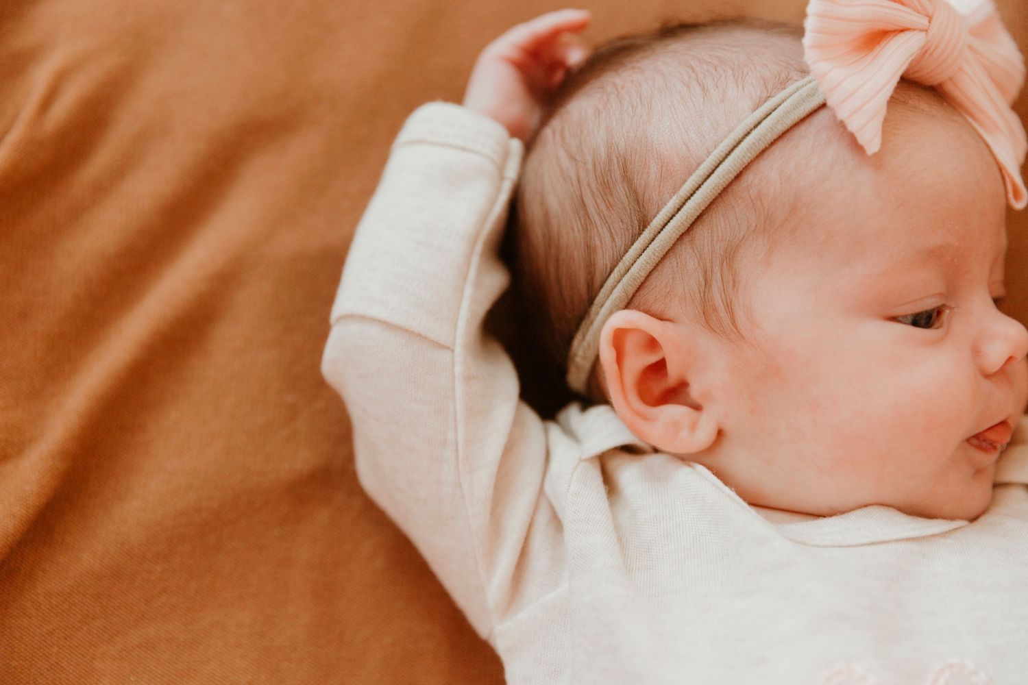 Close-up of sleeping baby in white outfit with pink bow on brown surface.