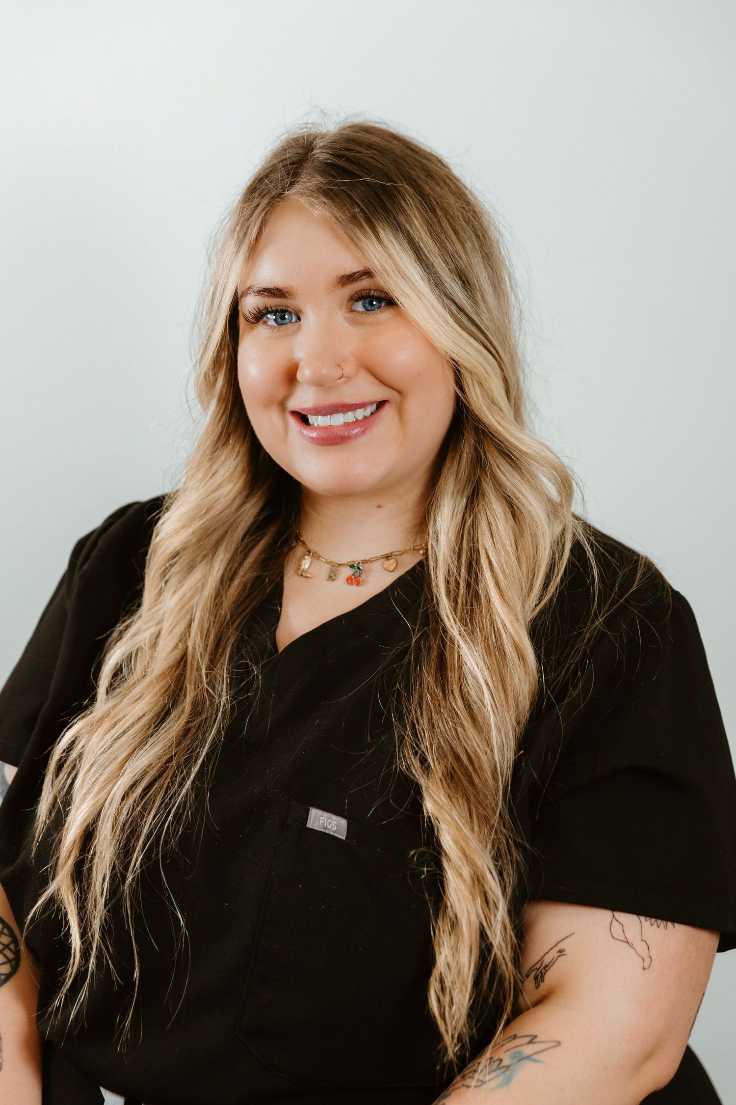 Professional headshot of someone in black scrubs with long blonde wavy hair and visible arm tattoos against light background.