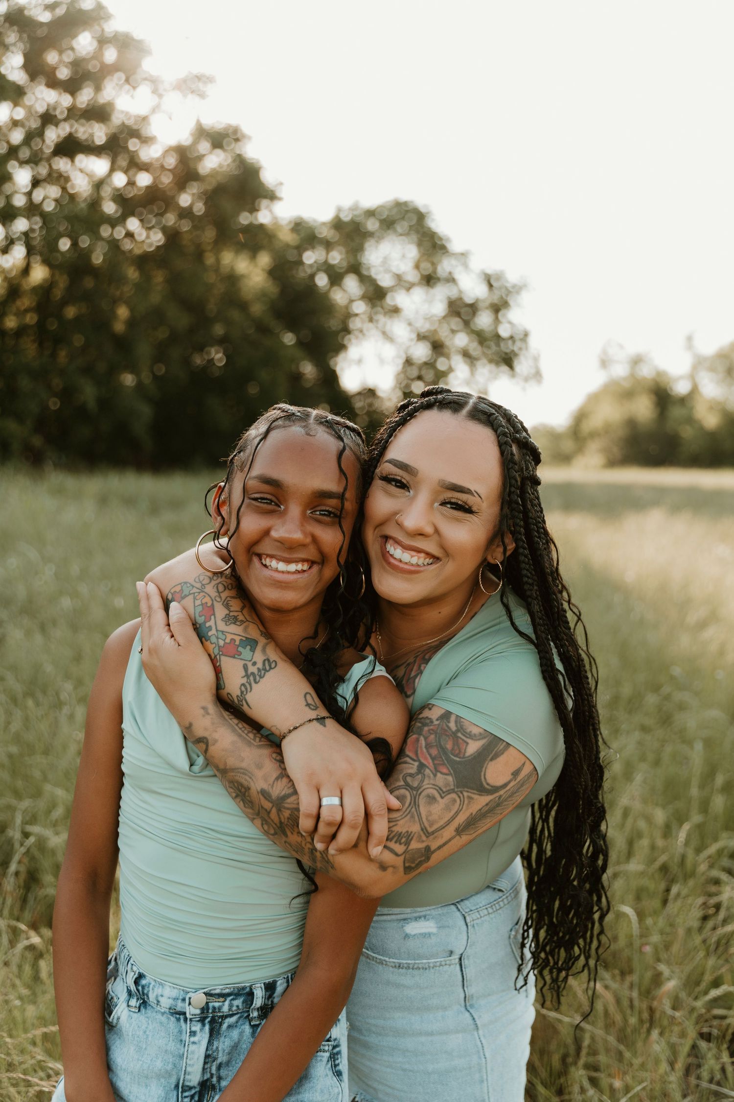 Two friends in matching mint green tops embrace and laugh in a sunny field.