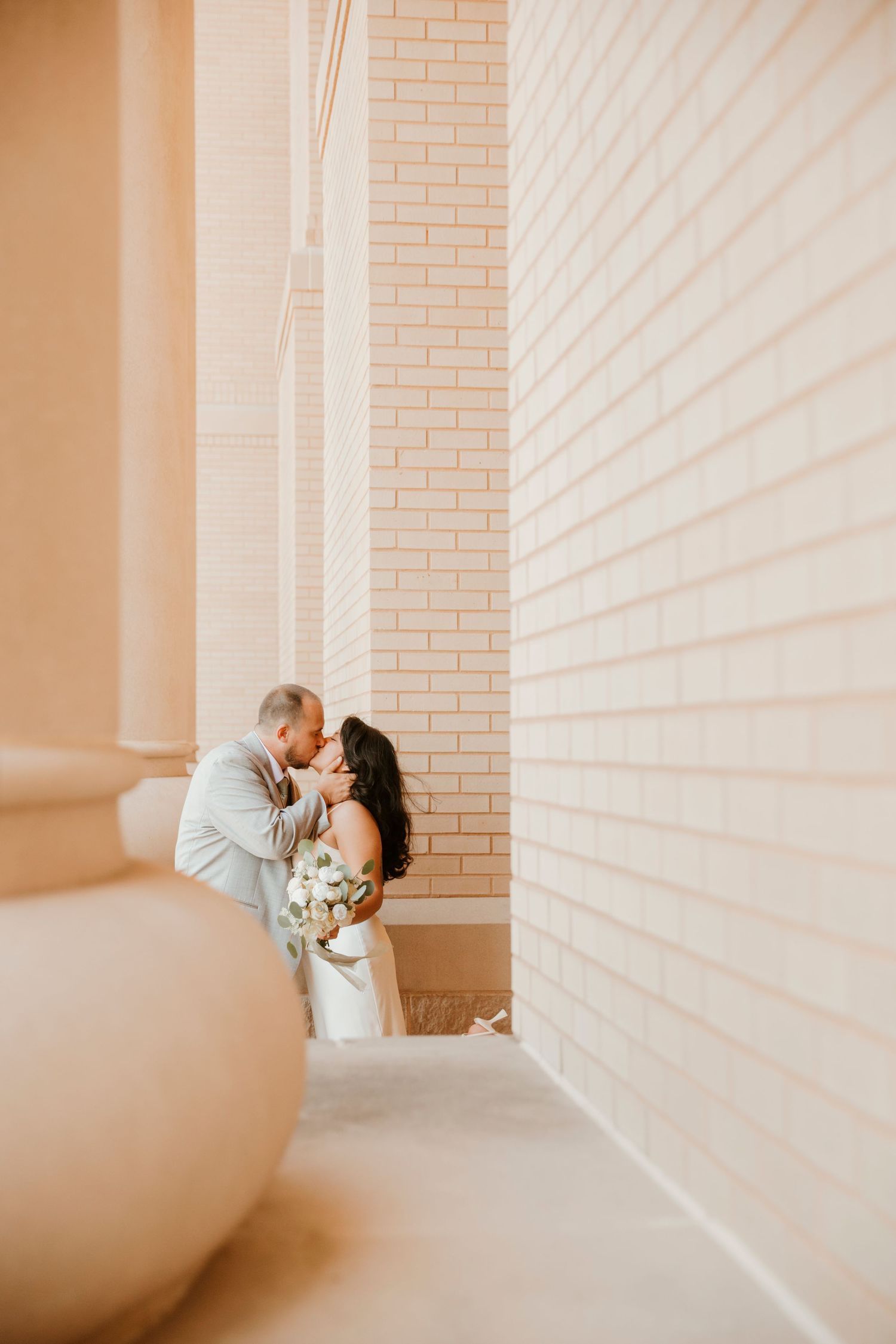 A romantic wedding moment captured in a light-filled corner with peach brick walls and soft natural lighting.