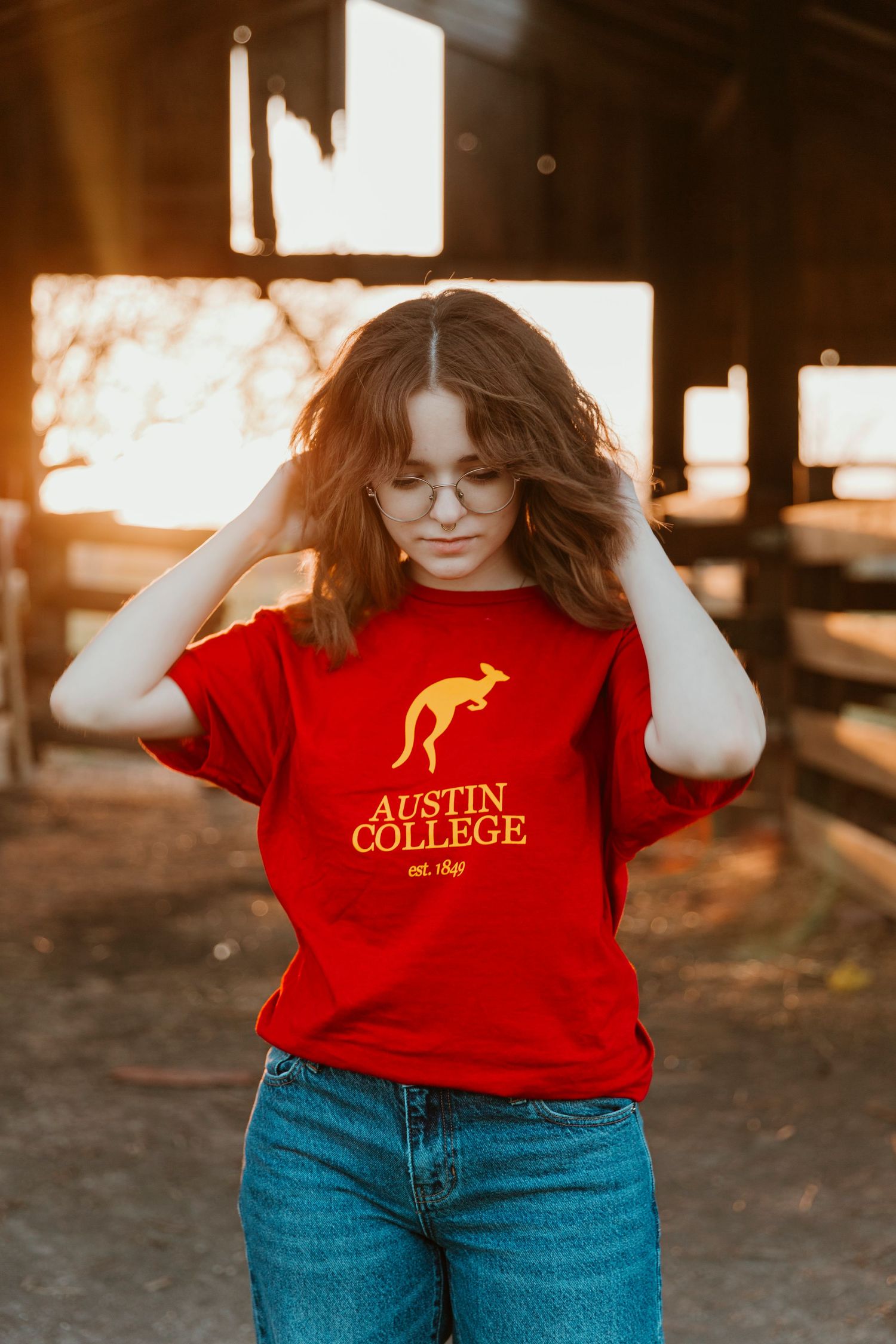 A red Austin College t-shirt worn with jeans in a rustic barn setting at sunset.