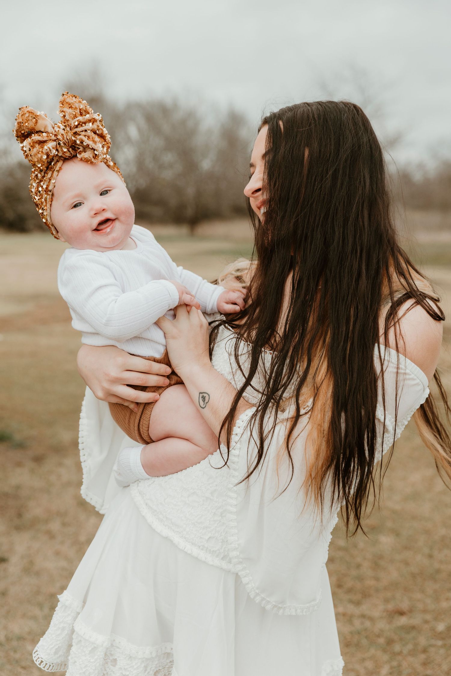 Mother in white dress holding smiling baby with orange bow headband outdoors.