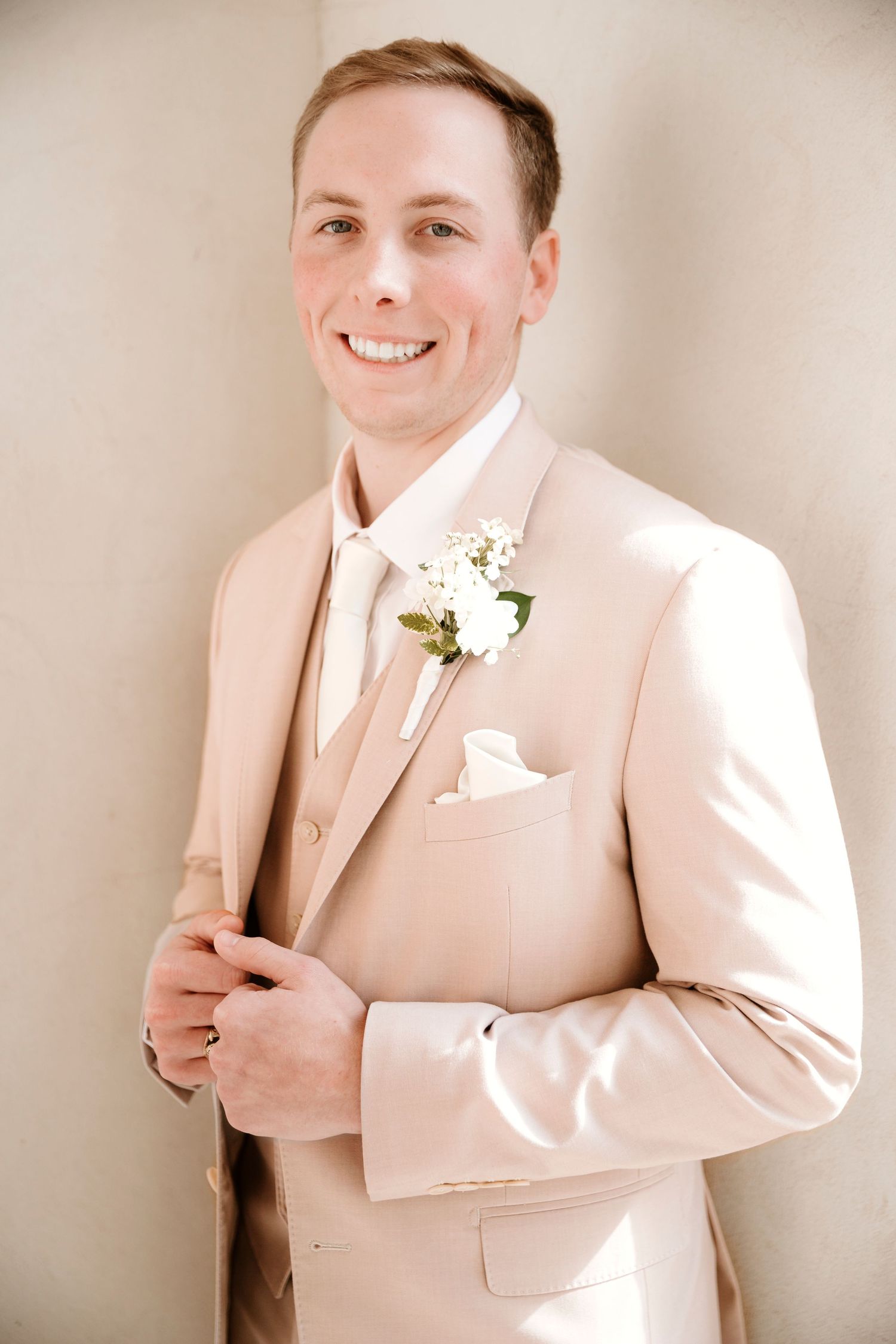 A person wearing a light beige suit jacket with a white boutonniere poses against a neutral background.