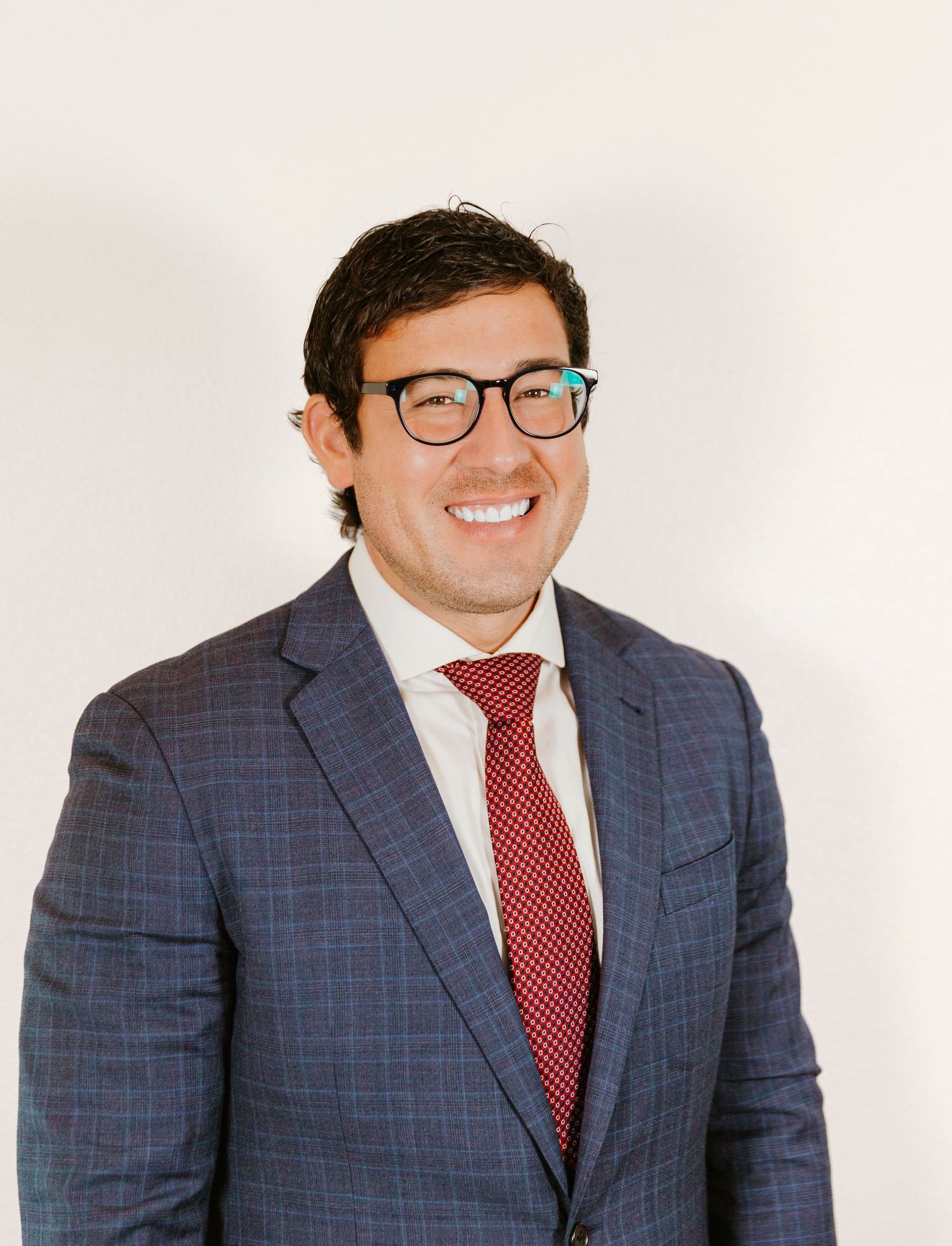 Professional portrait of person in plaid suit with red tie and glasses against white background.