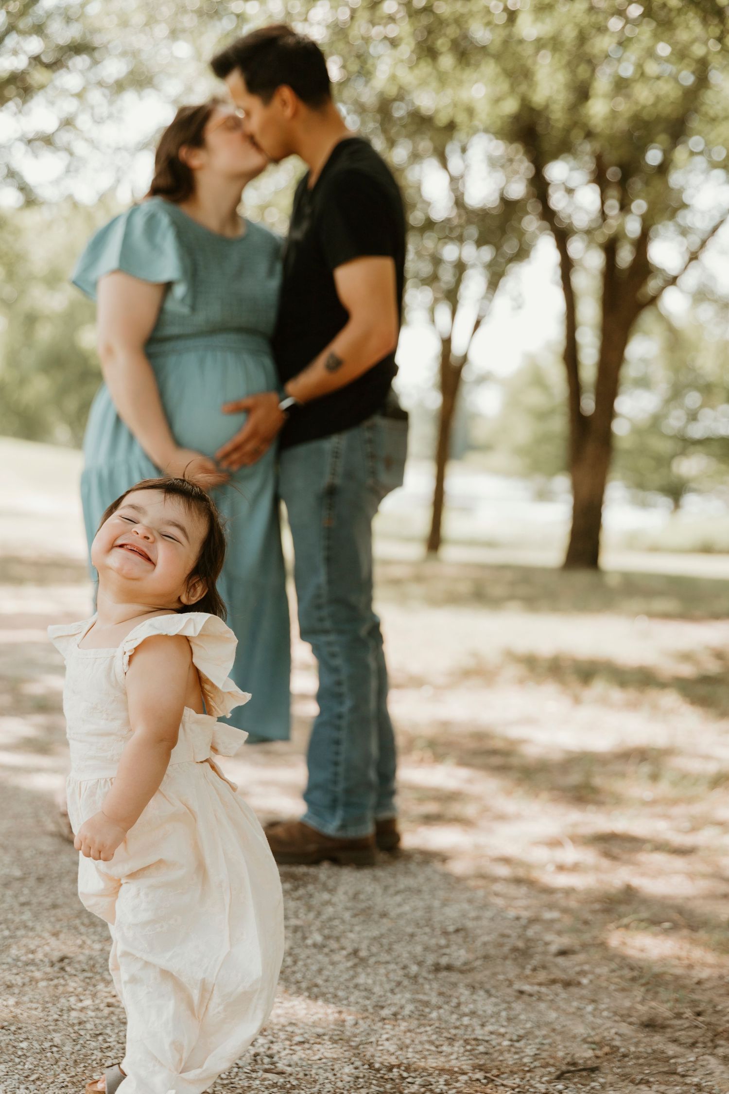 Sweet family moment in a park with a small child in white dress laughing in the foreground.