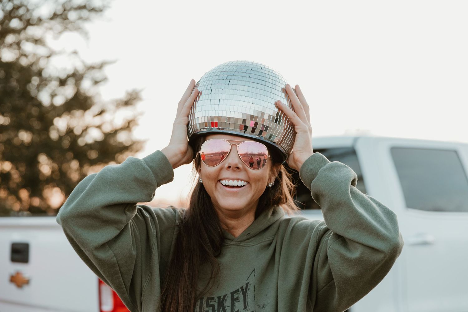 Person in green hoodie holds disco ball while posing against white camper background.