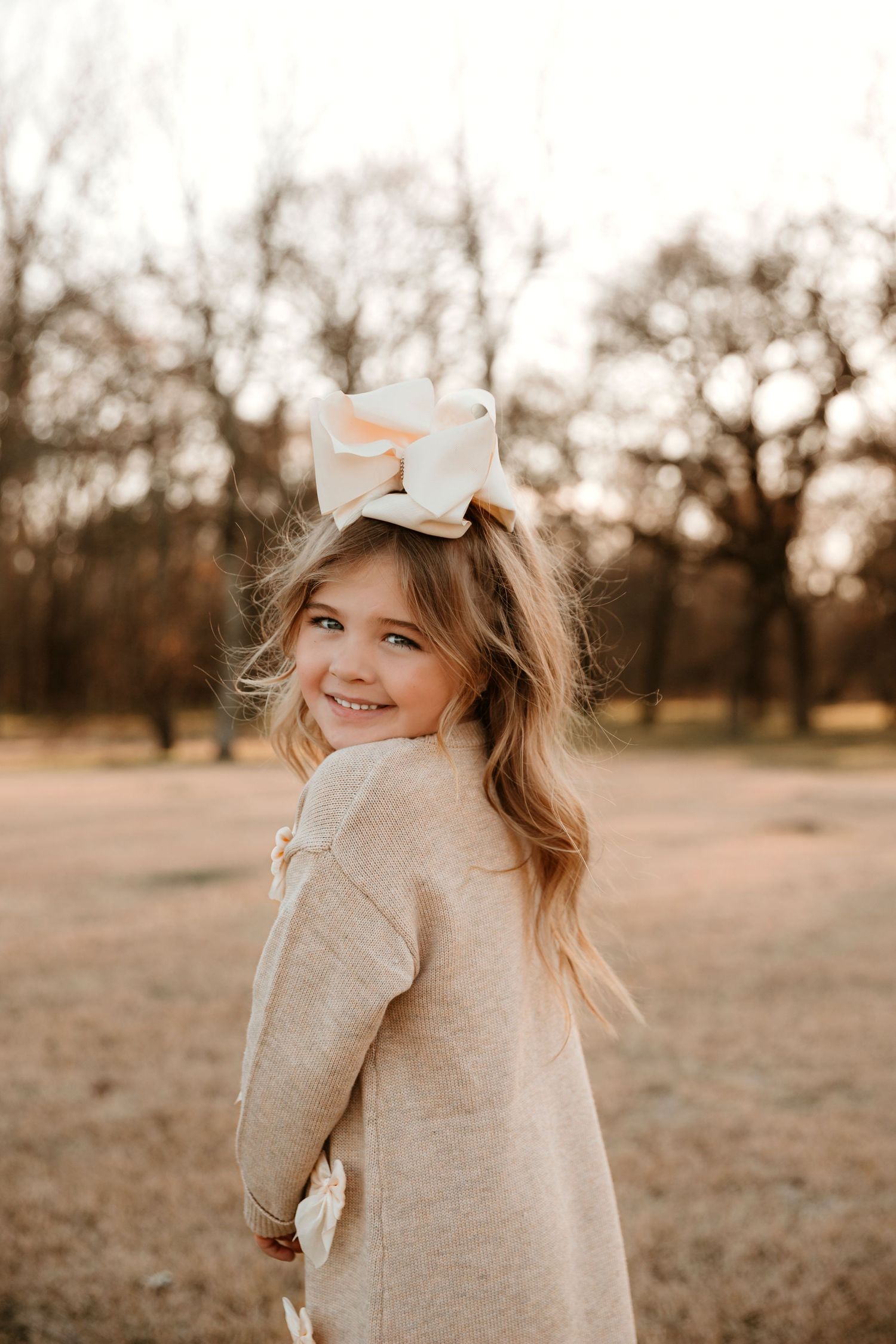 Young girl in beige sweater with bow in hair smiles over shoulder in autumn outdoor setting at sunset.