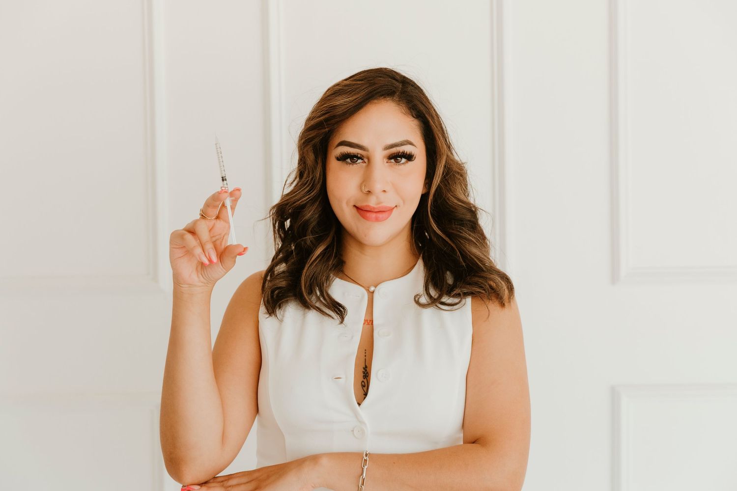 Person in white sleeveless top holds up finger while standing against white wall with soft smile.