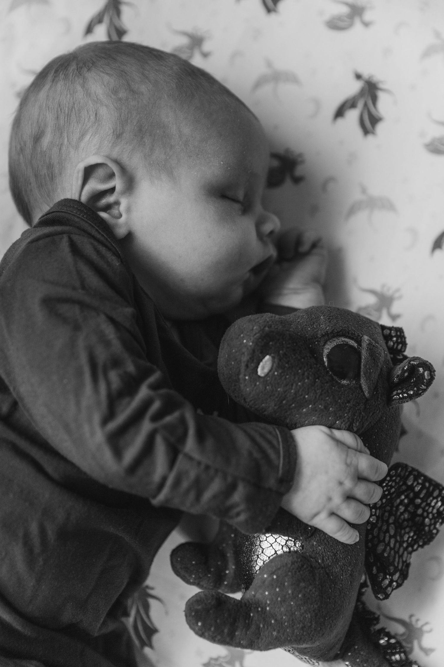 A sleeping child holds onto a plush dinosaur toy against a patterned wallpaper background in black and white.