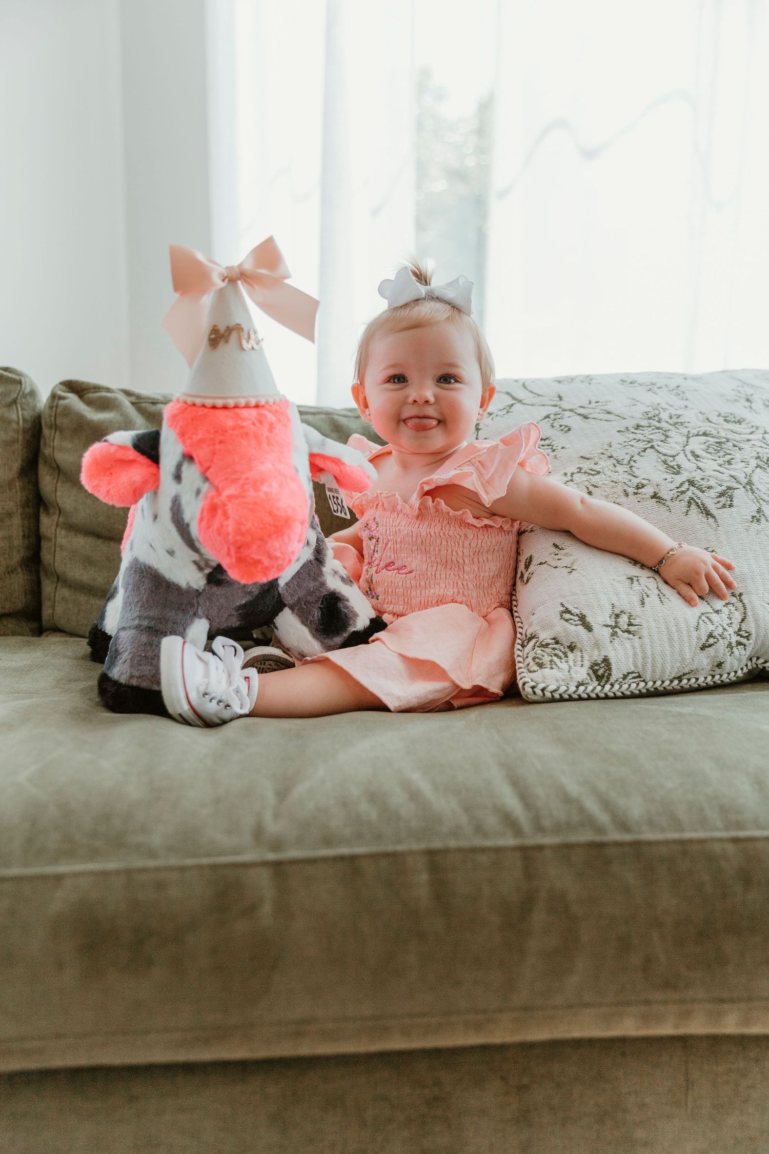 A smiling baby girl in a pink dress sits on a green sofa next to a stuffed animal wearing a party hat.