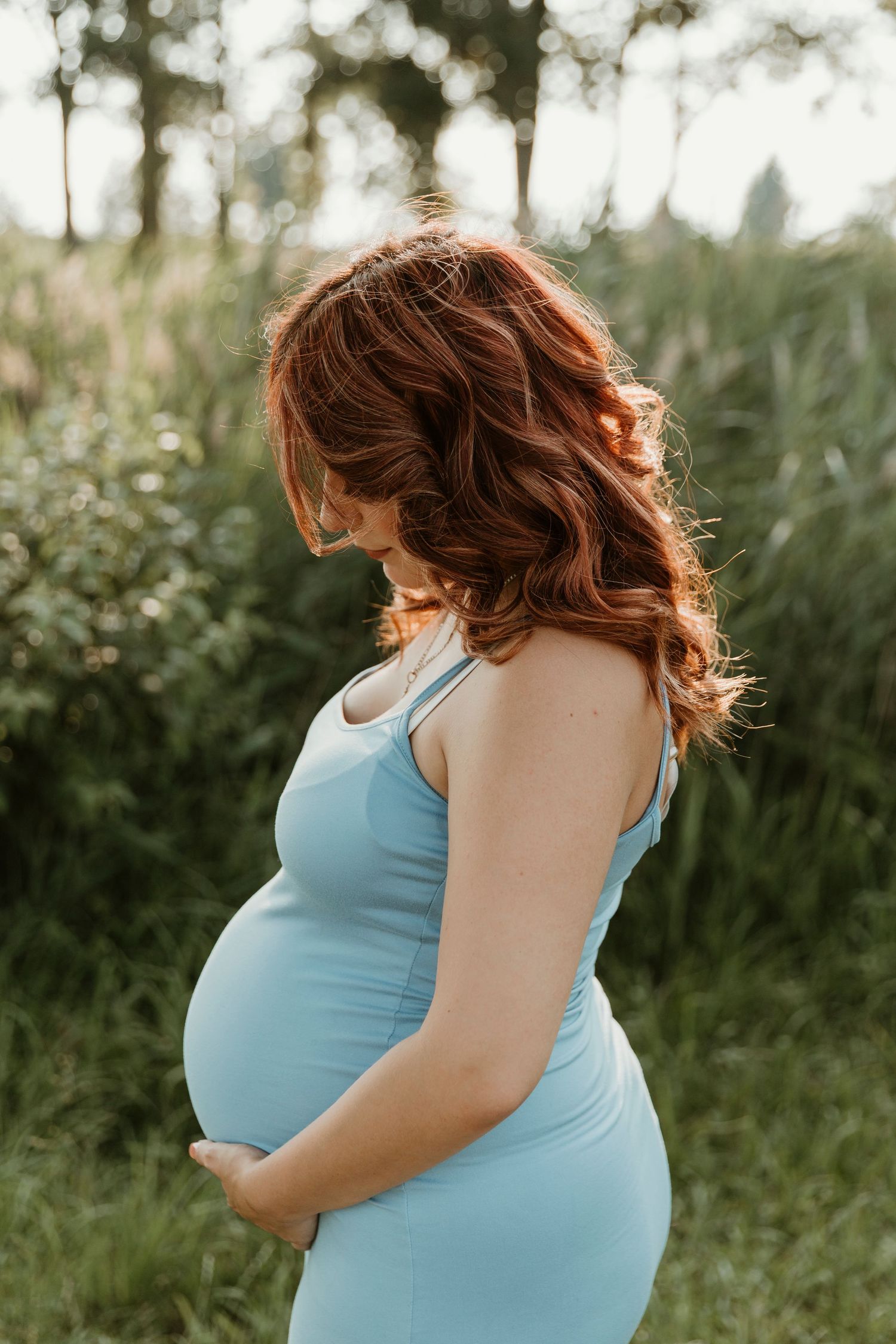 Side view of an expectant mother in a blue tank dress standing in a sunlit natural setting.