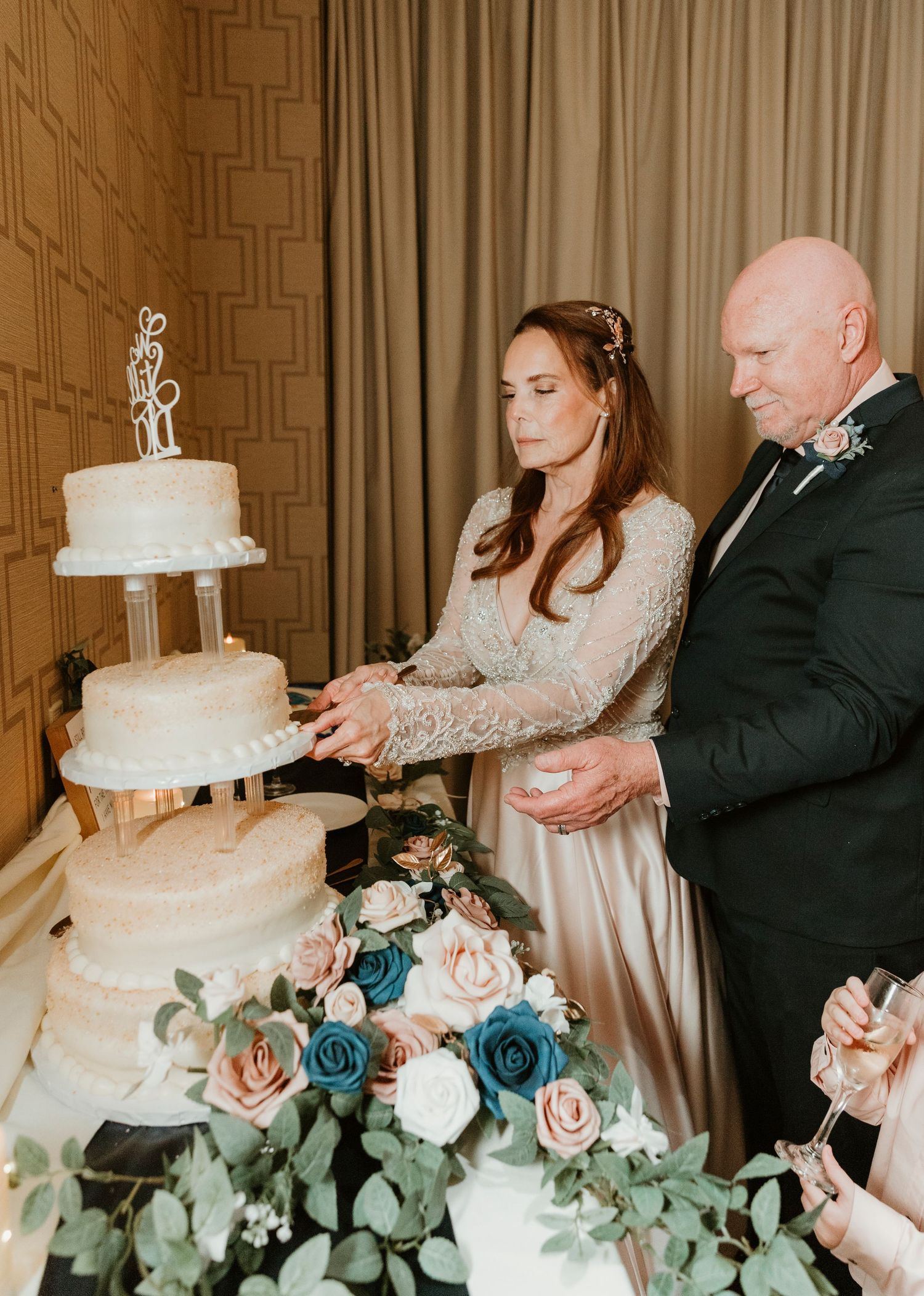 A couple cuts their elegant three-tier white wedding cake adorned with blue and pink roses and eucalyptus greenery.