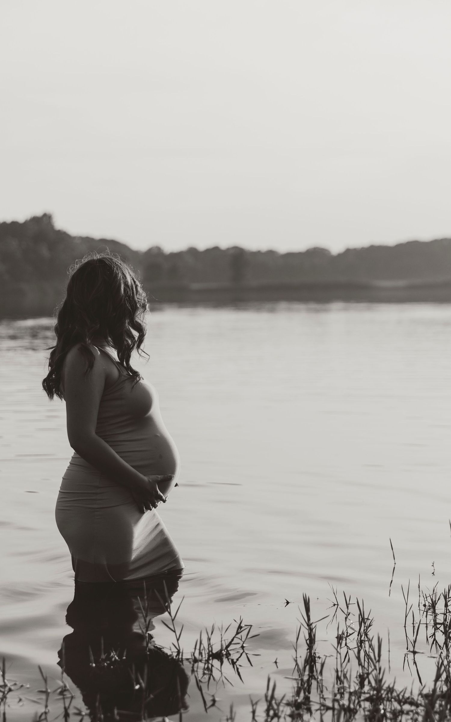 Black and white maternity portrait captured at the edge of a serene lake during sunset.