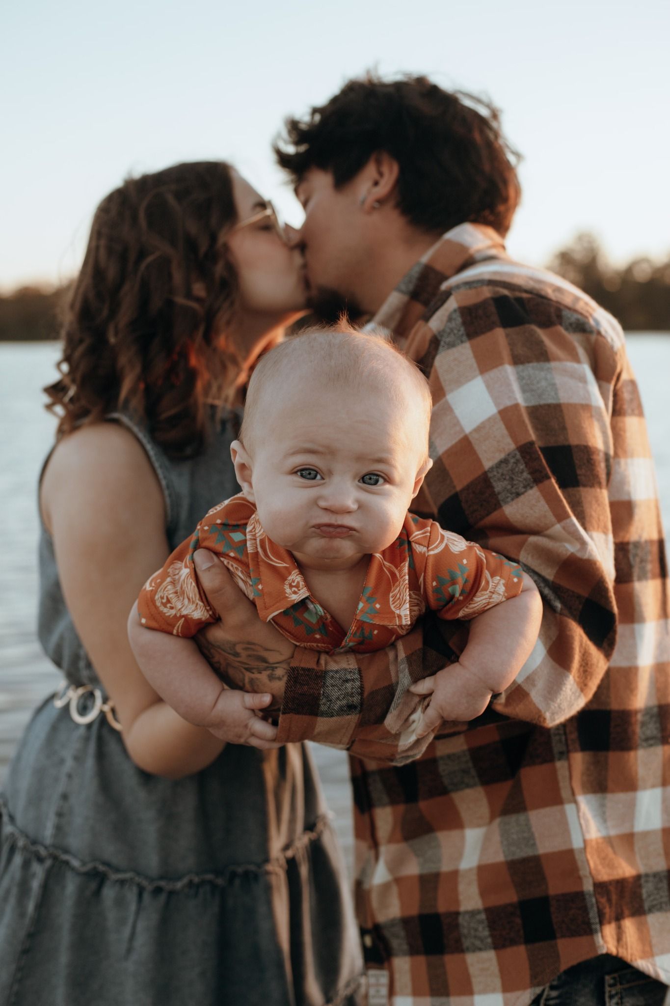 Sweet family moment captured by the water during sunset with baby in plaid shirt.