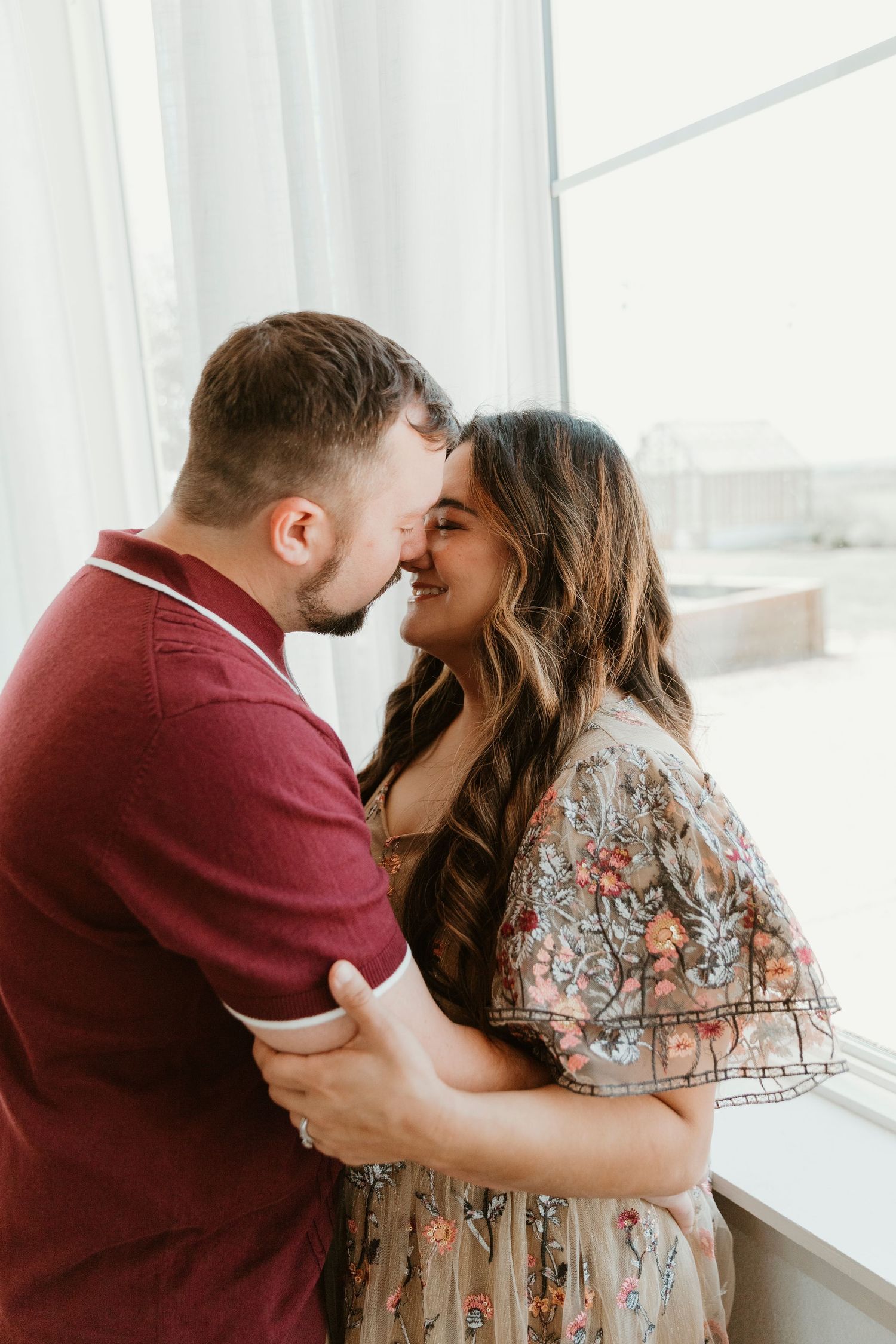 Intimate indoor portrait captures couple embracing near sunlit windows in modern setting.