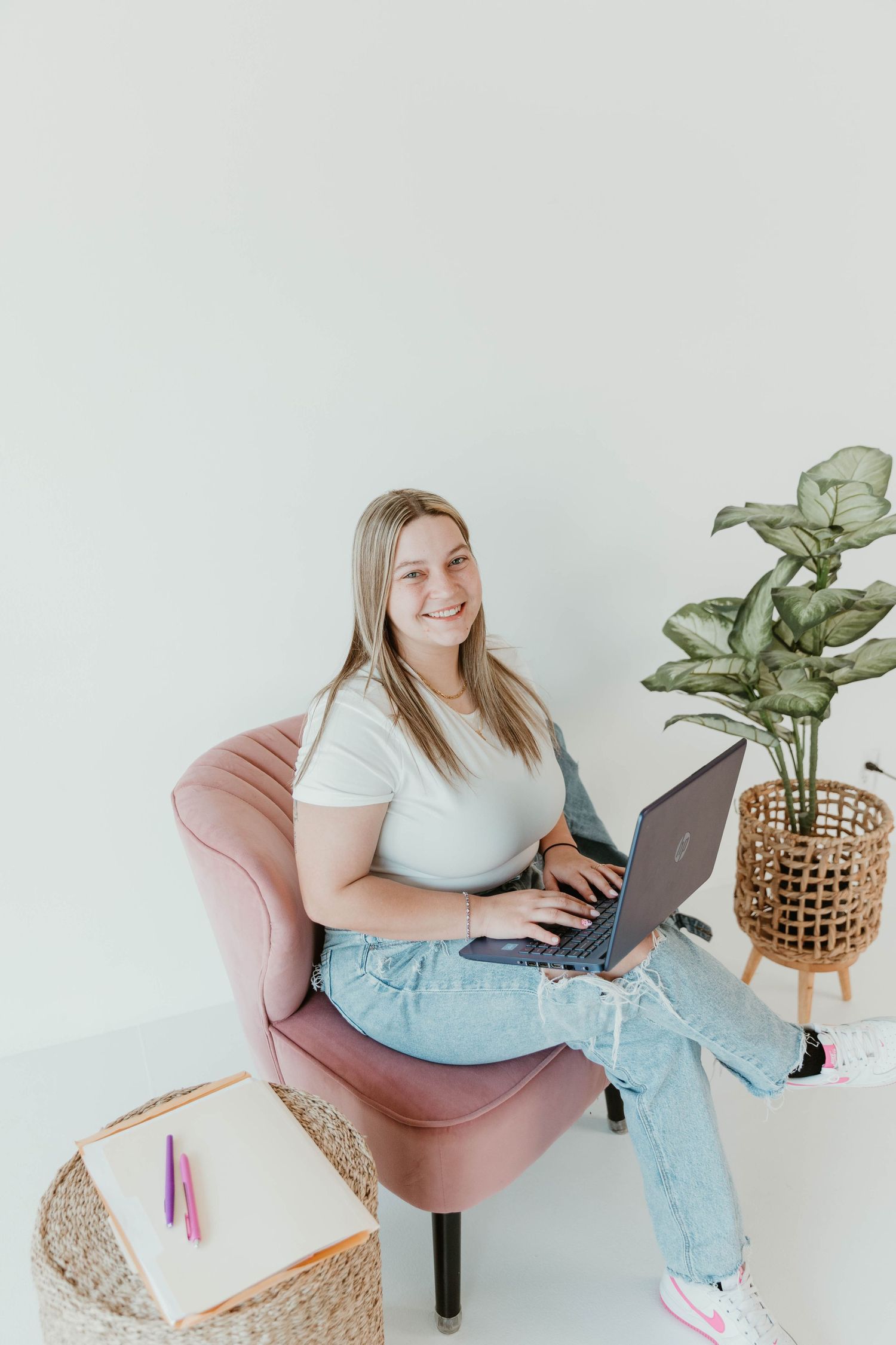 Person sitting in a pink accent chair with laptop and decorative plant in minimalist white room setting.