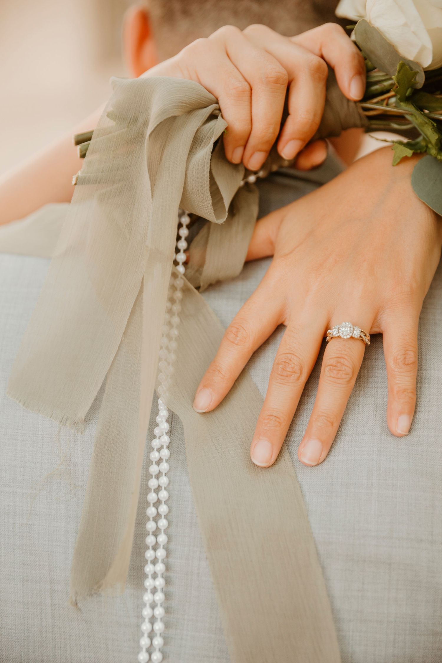 Close-up of a bride's hand showing engagement ring with pearl details and floral bouquet accents.