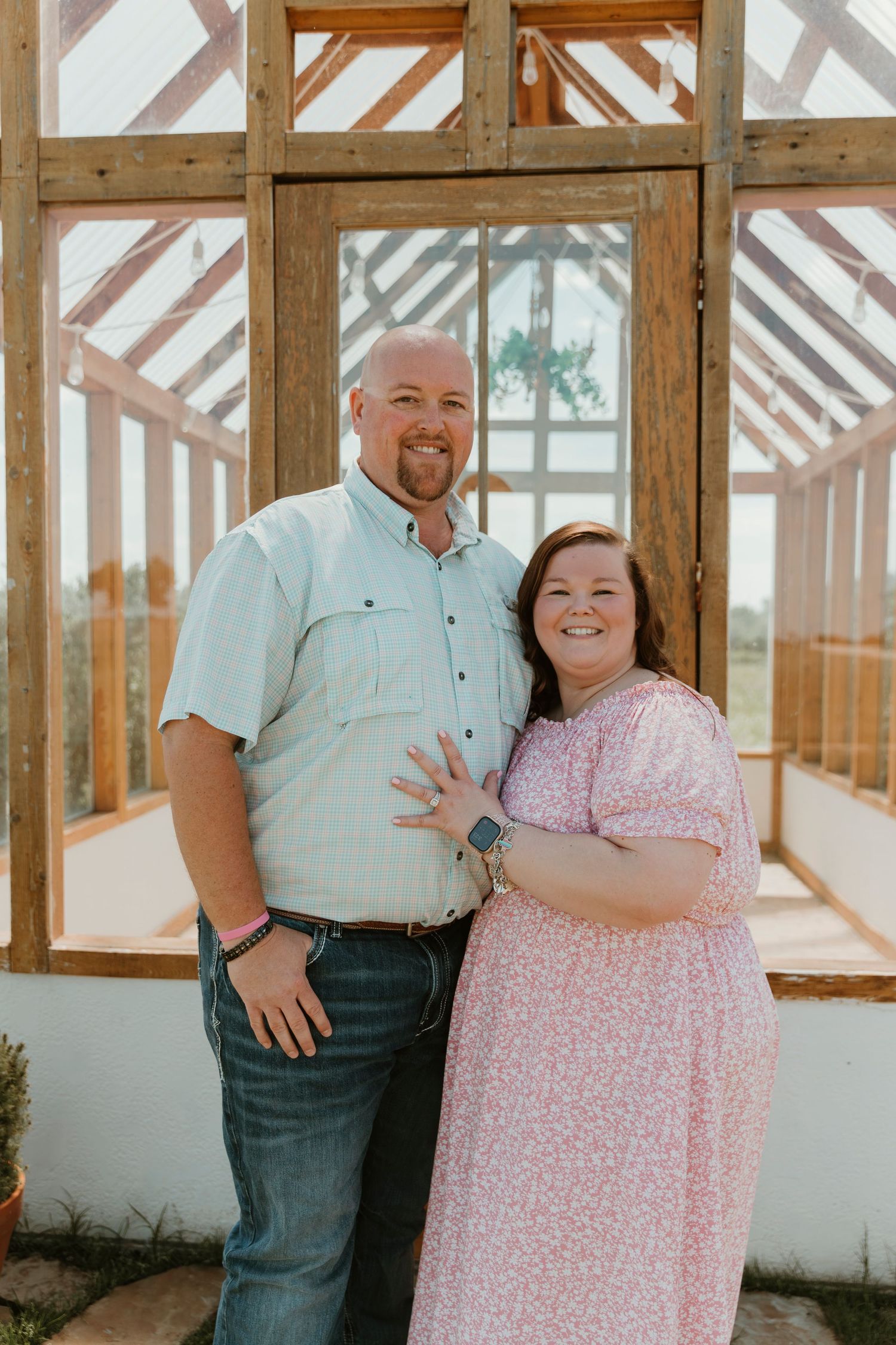 A smiling couple poses together in front of a wooden greenhouse, the woman wearing a pink floral dress showing off her ring.