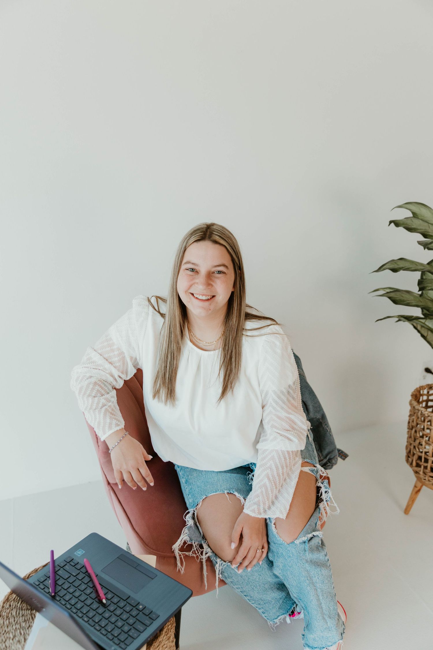 Person in casual attire sitting with laptop in minimalist white room with plant decor.