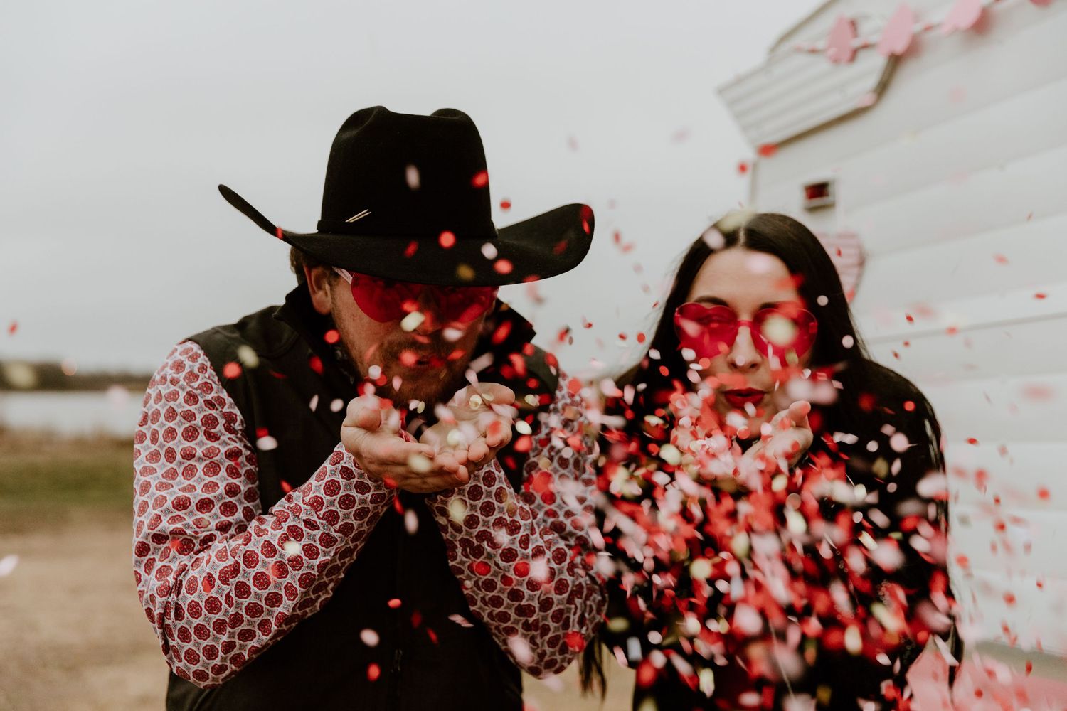 Playful couple blowing red confetti outdoors with one wearing a cowboy hat.