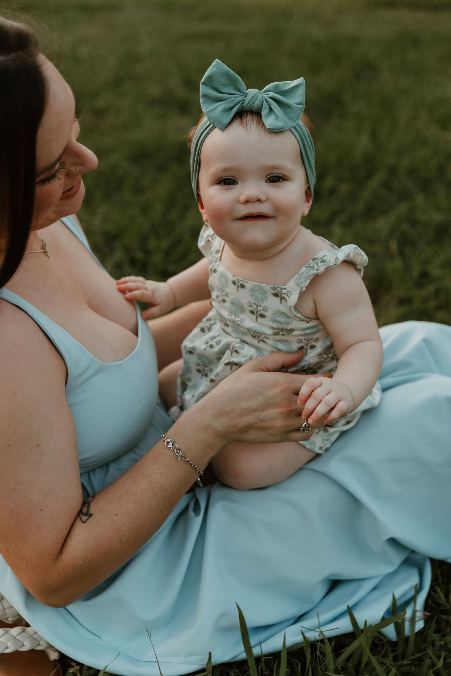 An adorable baby wearing a teal bow and floral outfit sits with someone in a blue dress.