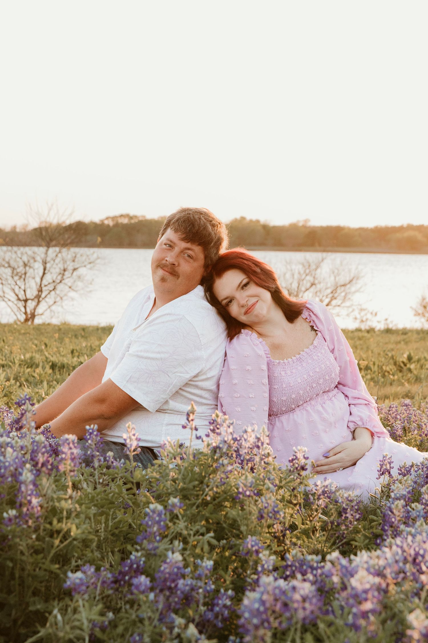 A couple sits together in a field of purple wildflowers at sunset near a lake.