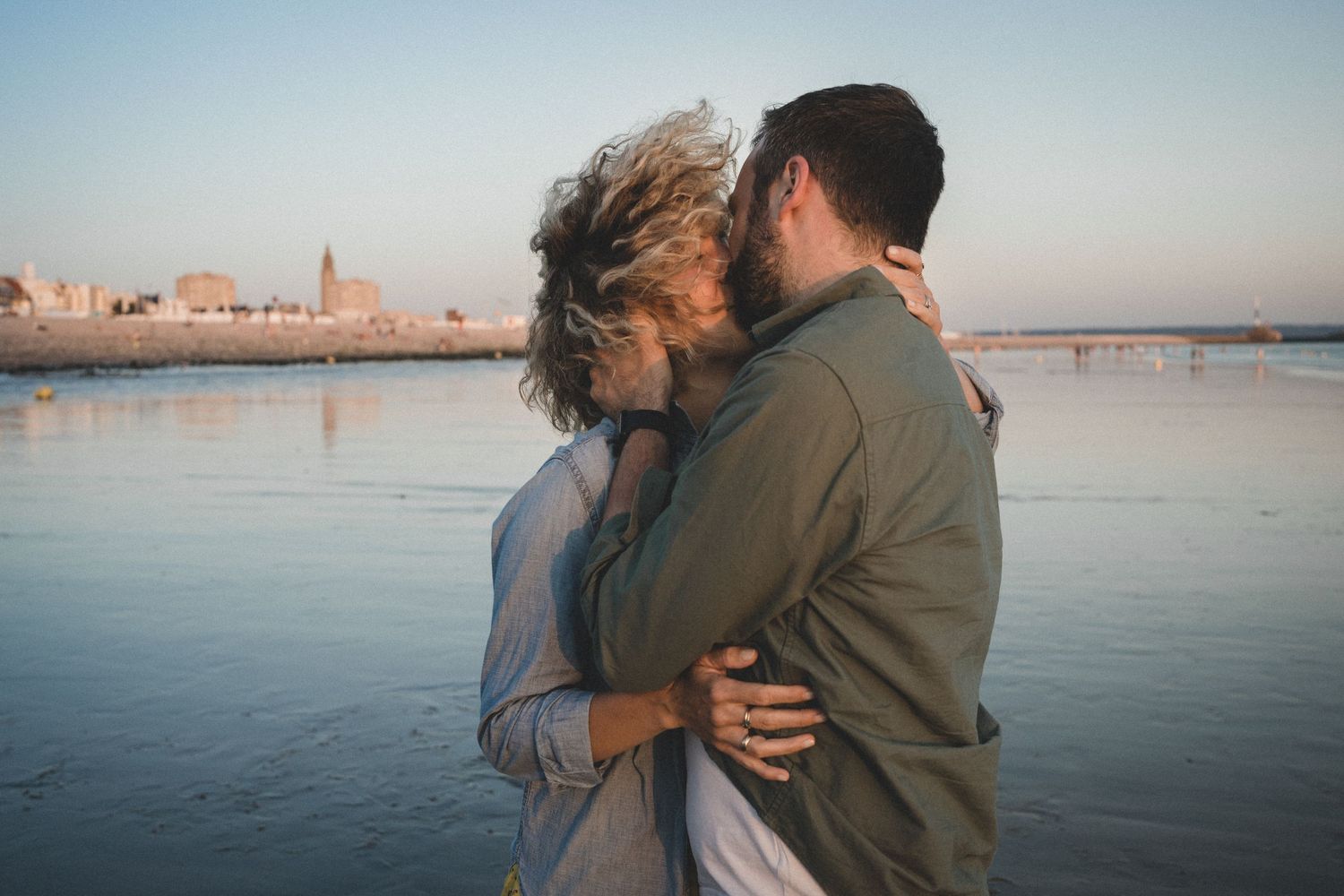 A couple embraces on a beach at sunset with a cityscape reflecting in the calm water.