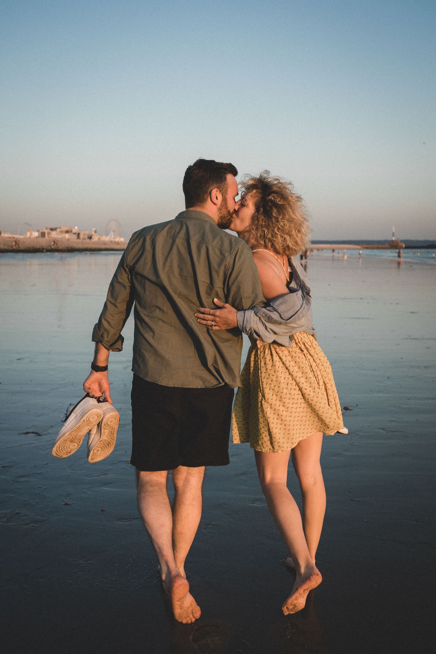 Couple sur la plage au coucher du soleil marche sur le sable au bord de l'eau en s'embrassant avec la ville du Havre en fond. Séance photo